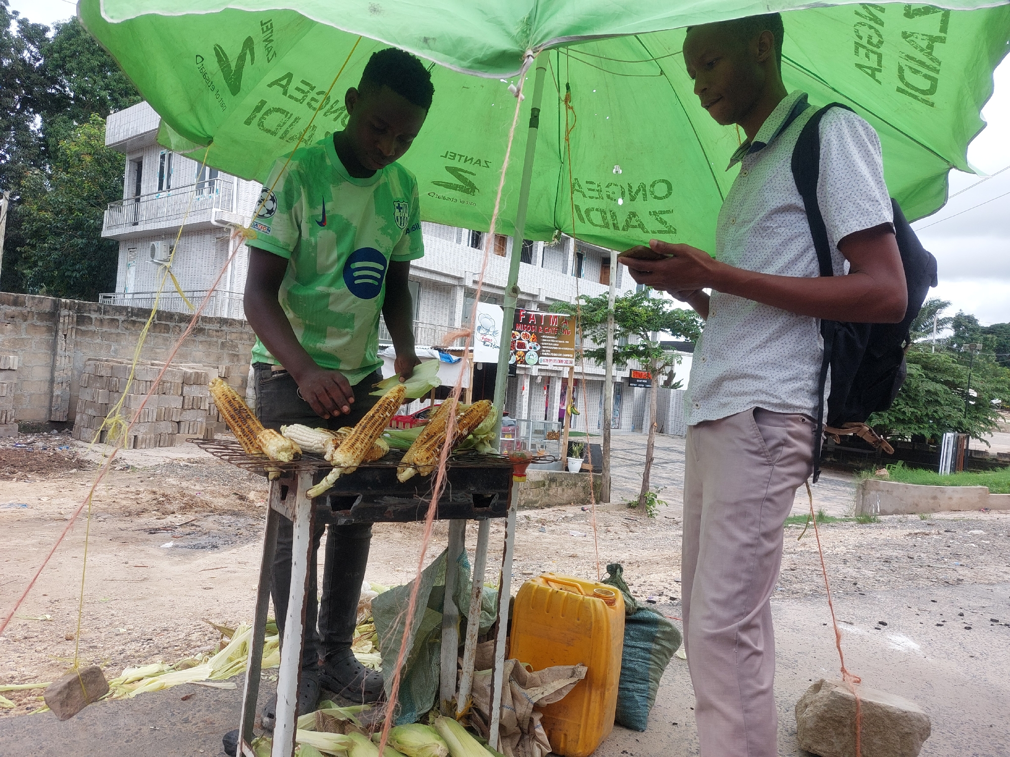The image depicts a street scene in Dar es-Salaam, Tanzania. A young man wearing a green patterned jersey is grilling corn on a makeshift grill stand. A customer stands nearby, looking at a mobile phone. Both are under a large green umbrella branded with logos and text, including the word "JETWAS". The street setting shows a partially constructed building in the background, along with other street-side establishments indicated by signage. A yellow jerrycan sits near the grill. Rocks are used to weigh down the umbrella's support ropes. The ground is unpaved.