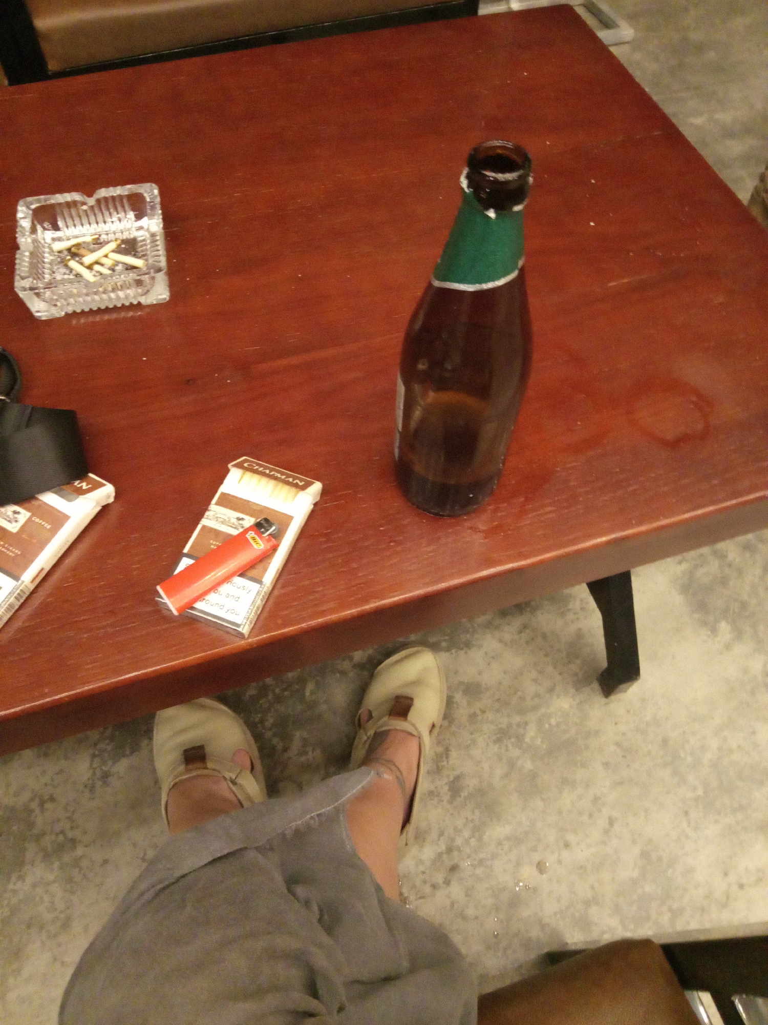 The image depicts a table in Nha Trang, Vietnam. On the table, from left to right, is a clear glass ashtray containing several cigarette butts, a partially visible black bag, two packs of Chapman cigarettes with a red lighter lying on top of one, and an open brown glass beer bottle with a green label covering its neck. A small puddle of liquid is visible to the right of the bottle. Below the table are two feet wearing tan sandals and part of a light grey garment. The floor appears to be concrete.