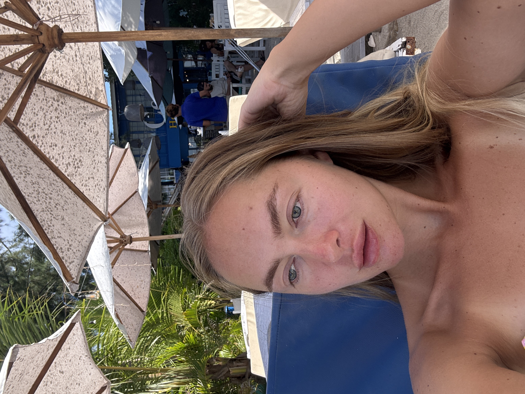 A selfie shows a woman lying on a blue lounge chair. Beach umbrellas provide shade overhead. Green palm trees and other tropical vegetation are visible in the background, along with a building structure and people. The photo appears to be taken at a beach location, likely in Unknown, Indonesia.