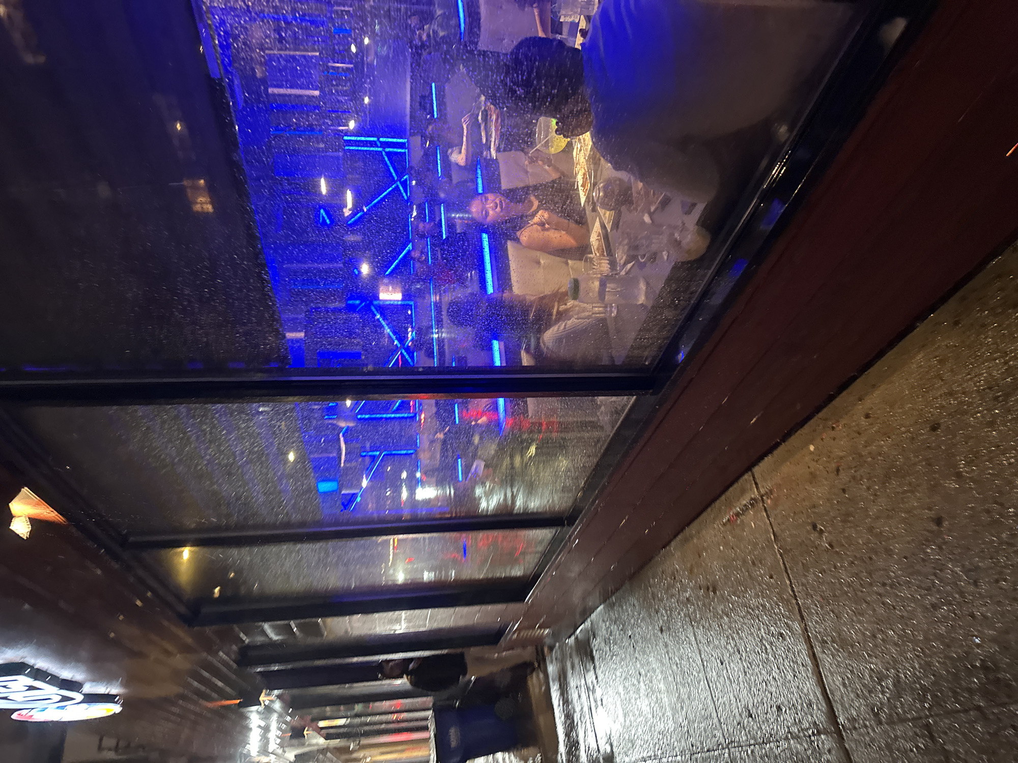 The image depicts a rainy evening scene in New York City, United States of America, as viewed from under an awning. Raindrops streak across the glass of the awning, obscuring and distorting the view. Reflected in the glass is the interior of what appears to be a restaurant or bar, illuminated with vibrant blue and red neon lights. Several people are seated at tables inside, appearing to be engaged in conversation and dining. Outside, the wet pavement reflects the ambient light, creating a shimmering effect. A few pedestrians can be seen walking along the sidewalk. The overall atmosphere is one of urban activity on a wet and somewhat dreary night.