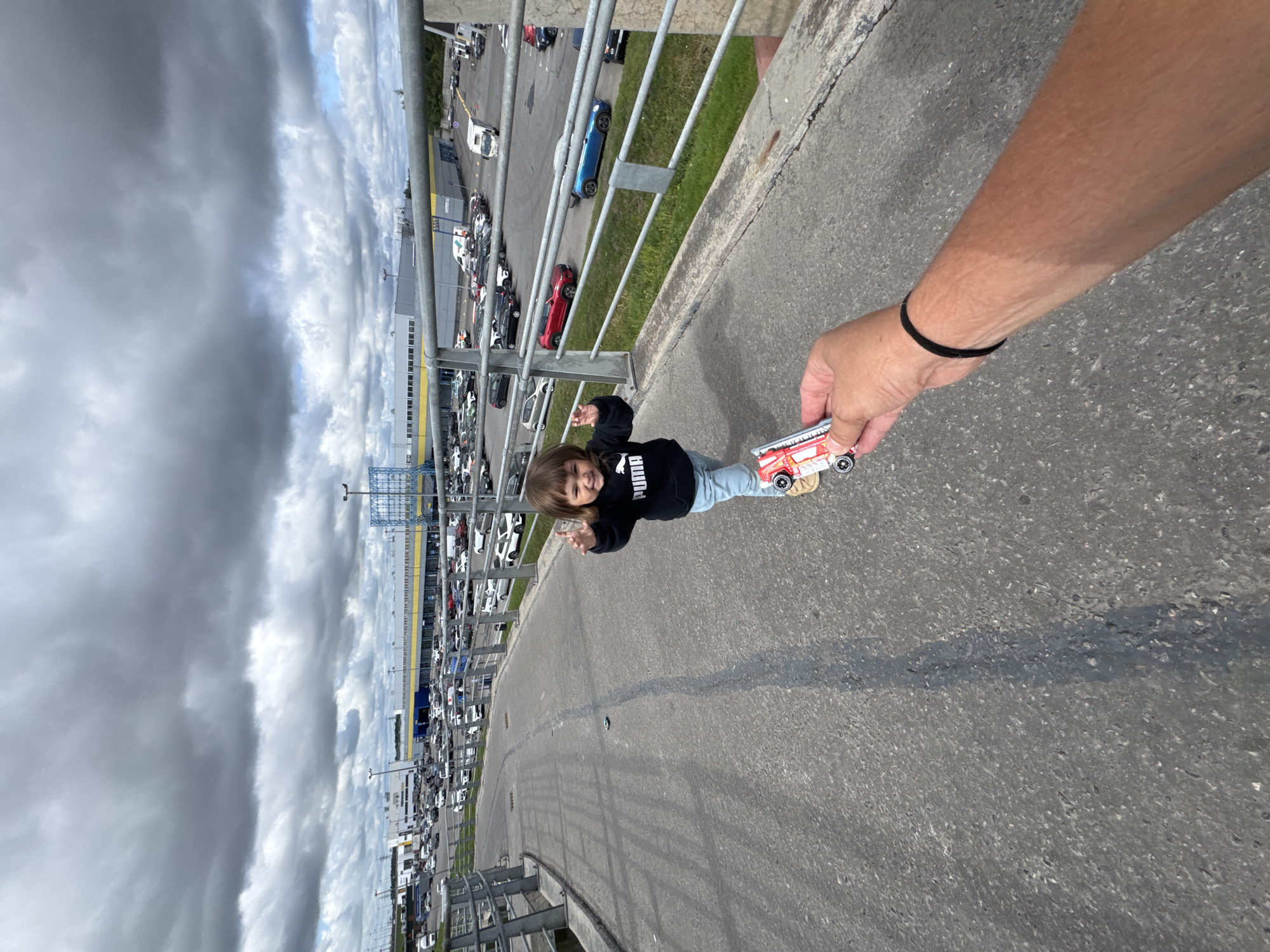 A young child, a girl, stands on an asphalt path, holding a toy car. She is wearing a black Puma hoodie and blue jeans. Her left arm is raised, and her hand is open, with her thumb and index finger extended. She is smiling and looking towards the camera. The path is a pedestrian walkway, possibly a bridge or overpass, with a metal railing on the right side. In the background, a large parking lot is filled with cars, and a long, low-rise building with a yellow and blue facade is visible. The sky is overcast with dark, dramatic clouds. The scene appears to be in a commercial or industrial area of Saint Petersburg, Russia, likely near a shopping center or other large retail establishment, given the extensive parking lot and building architecture. A person's arm and hand are visible in the foreground, holding another toy car, suggesting an interaction between the person and the child. The asphalt path has a dark, painted line running down the center. The presence of the toy car in the foreground and the child's engagement with a toy further indicate a playful or recreational context within this urban environment.
