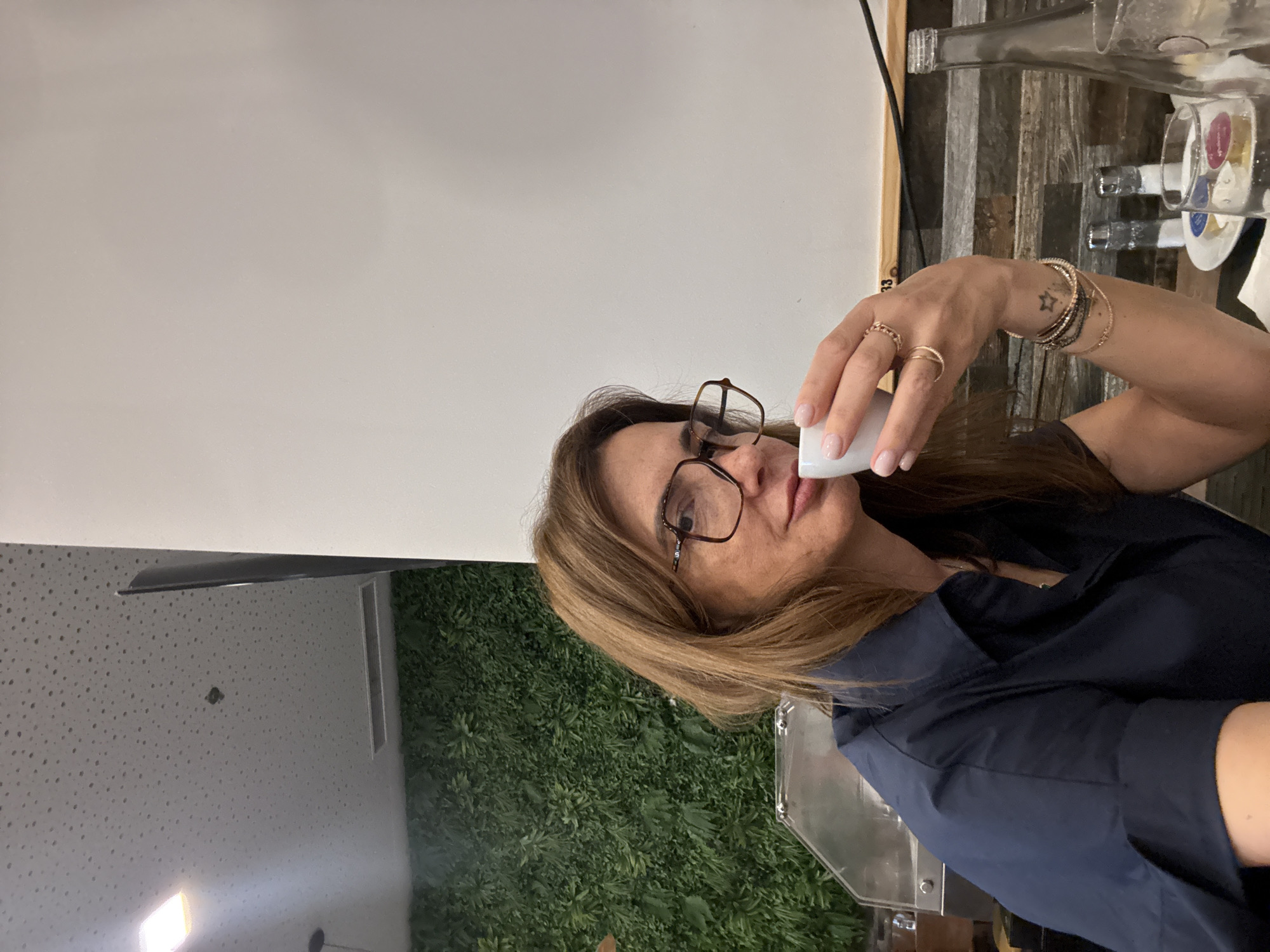 The image depicts a woman in Odivelas, Portugal, drinking from a small white cup. She wears glasses and a navy blue shirt. Her left wrist has multiple bracelets and rings. In the background, there is a white wall, a green wall with plant decorations, and a partially visible table with wine bottles and glasses. The location is likely a restaurant or bar in Odivelas, given the setting.
