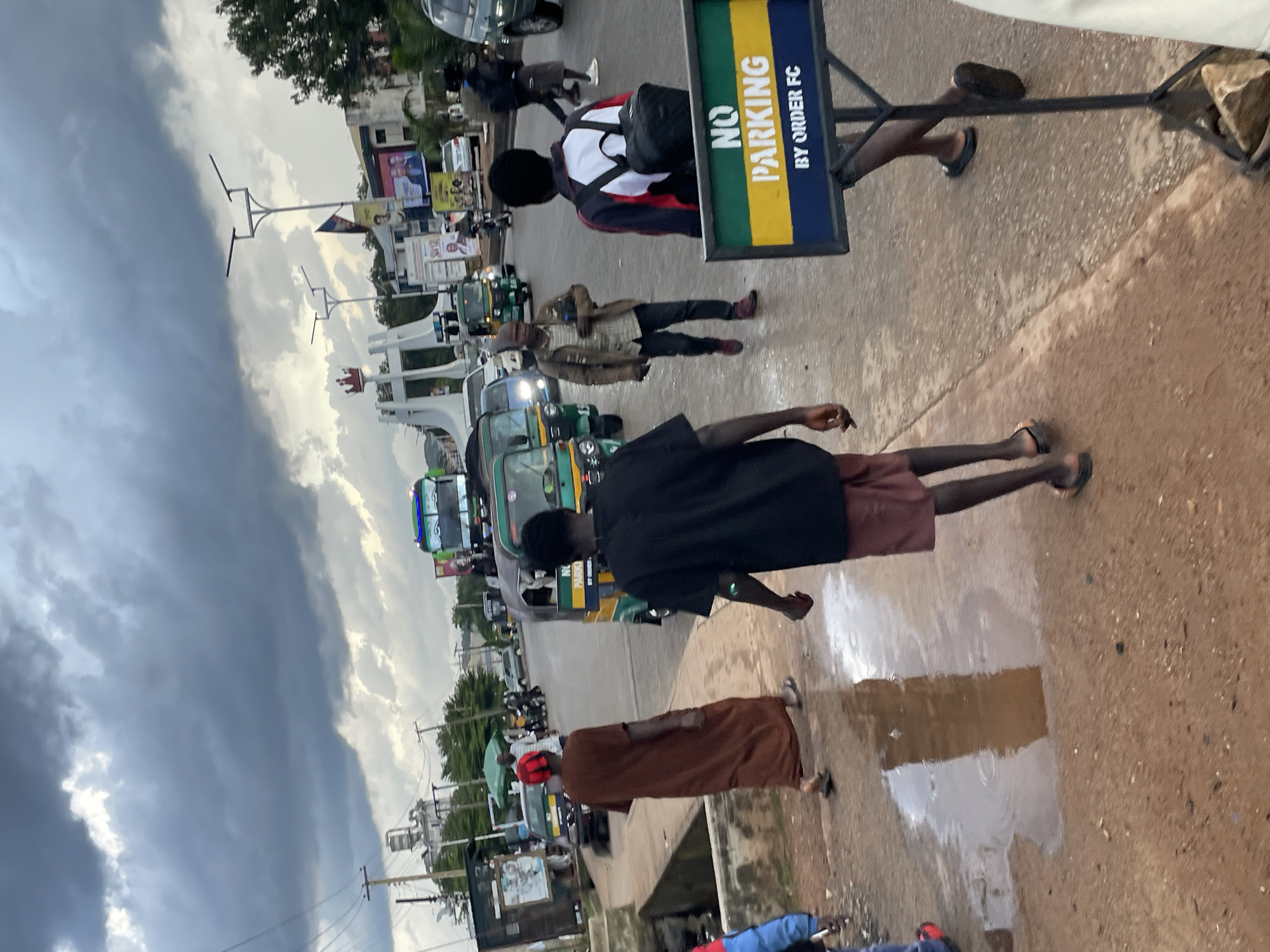 The image depicts a street scene in Jos, Nigeria, under a cloudy sky. In the foreground, a person wearing a brown robe walks on a wet, reddish-brown dirt road, with puddles reflecting the surroundings. To the right, another person in a black shirt and red shorts walks away from the camera, also on the wet road. A "NO PARKING BY ORDER FC" sign, with green, yellow, and blue stripes, stands on a metal frame. In the mid-ground, several auto-rickshaws, a green truck, and other vehicles are visible on a paved road. A person in a light-colored jacket and dark pants walks towards the right. In the background, a large white sculpture resembling a crown on a pedestal stands near a busy intersection with traffic lights and overhead wires. Billboards with political advertisements are also present. The overall atmosphere suggests a potentially rainy or recently rained-on day in a bustling urban area.