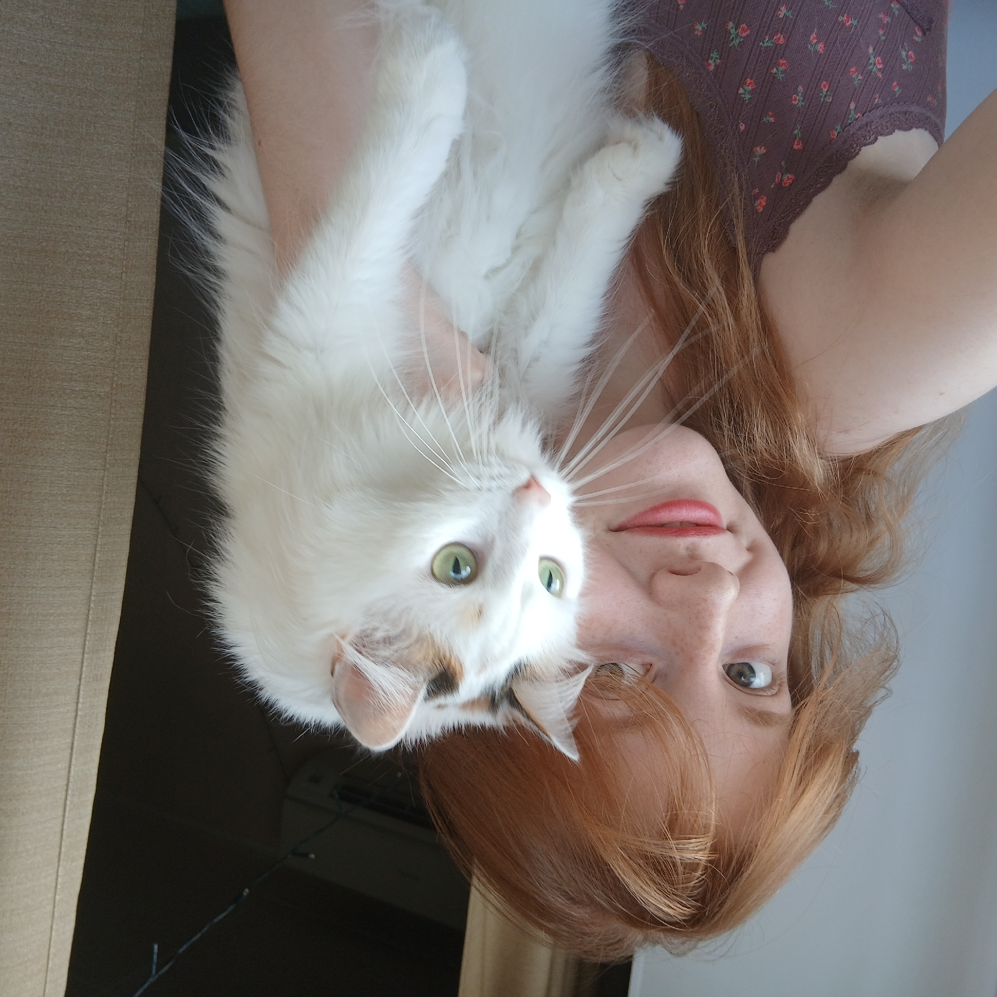 A woman with red hair and freckles is holding a long-haired white cat with green eyes. The woman is wearing a brown floral-patterned top. The cat is looking up and to the left. The background is blurred, but includes a beige wall and a dark area with Christmas lights hanging. There is nothing to indicate that the image was taken in Yekaterinburg, Russia.