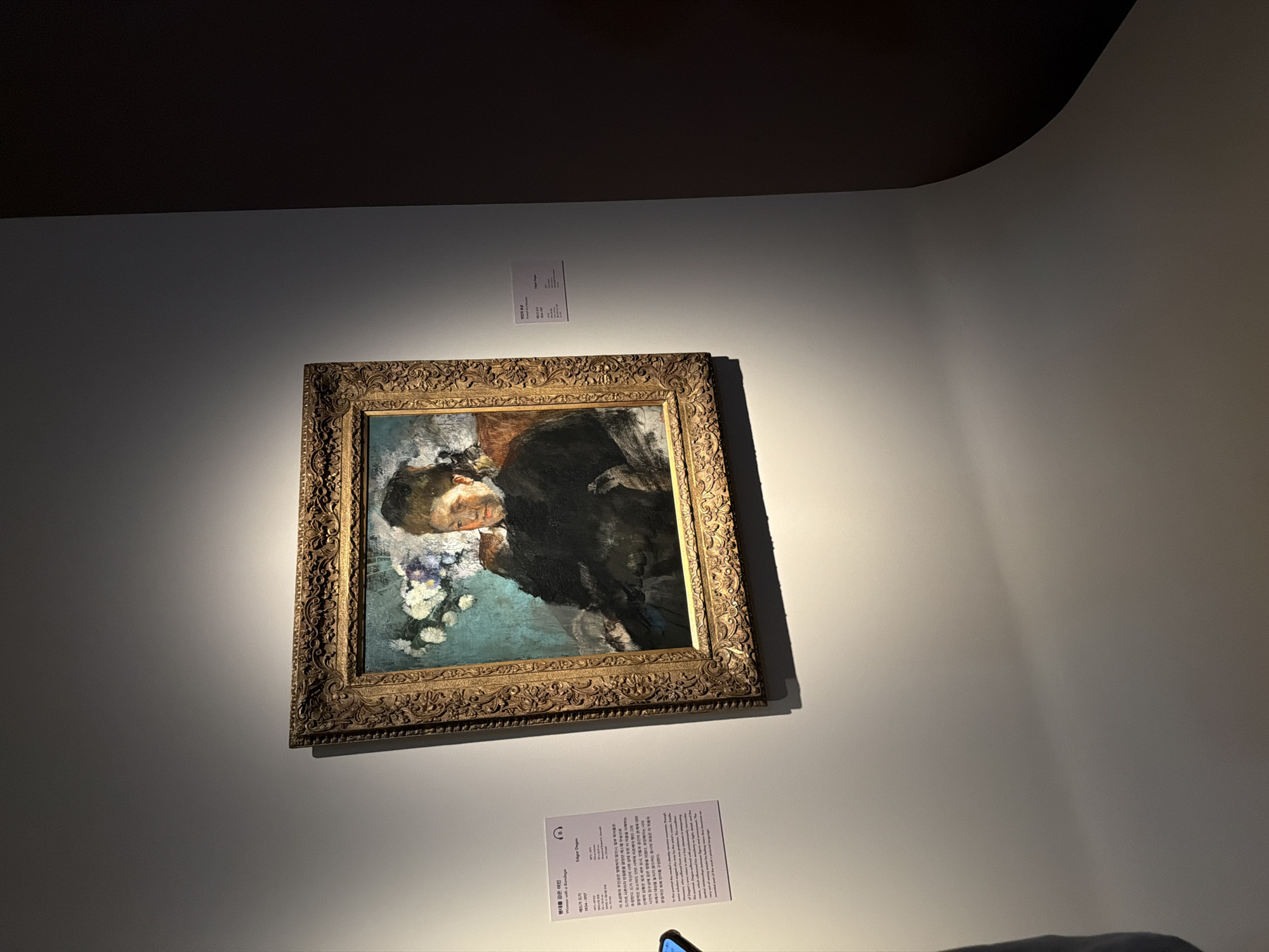 The image shows a painting of a woman in a dark dress with a dark background and a floral element above her head. The painting is in a gold ornate frame. To the left of the painting is a white label with Korean text and an English translation, identifying the artwork as "Woman with a Bondage" by Edgar Degas. The painting is displayed on a light gray wall, which is illuminated by a spotlight from above. The wall curves to the right into a darker background, suggesting the painting is in a museum or gallery. A phone is visible in the lower left corner, held by an unseen person, potentially taking a picture of the artwork. The scene appears to be indoors, in a dimly lit exhibition space.