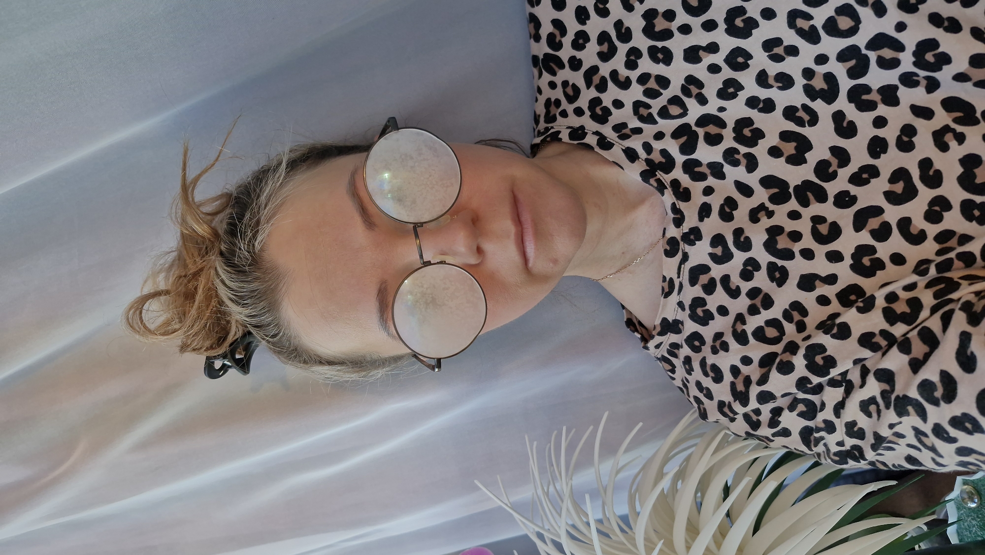 A fair-skinned person wearing round, wire-rimmed glasses is lying down on a surface of light blue fabric. The person has light blonde hair that is styled in a messy bun, held in place by a black claw clip. They are wearing a tan-colored shirt with a black leopard print pattern, and a thin gold necklace is visible around their neck. To the bottom right of the person is a white, palm-like plant. The setting is unknown.