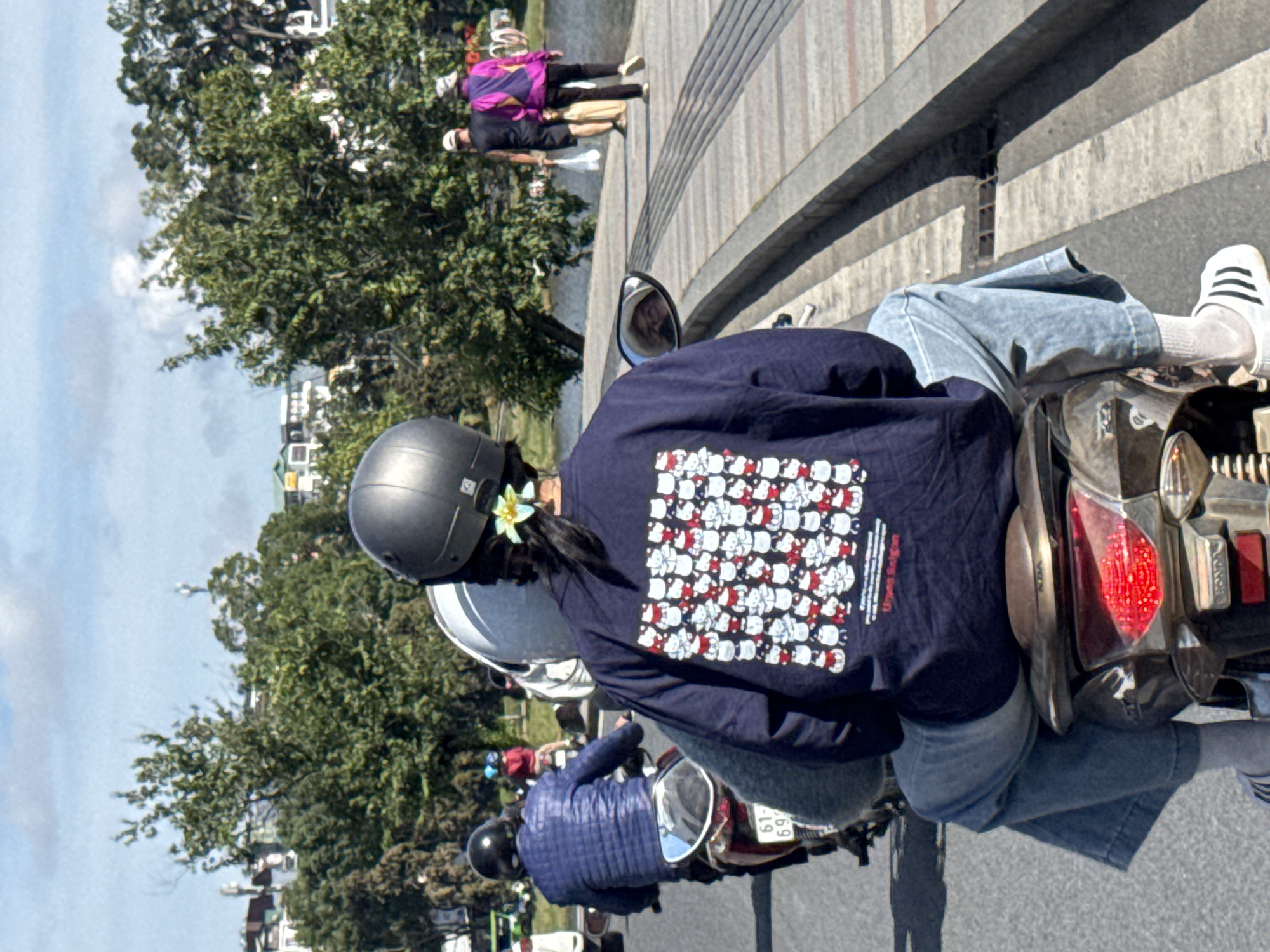 In Da Lat, Vietnam, on a bright, sunny day, a person wearing a navy blue sweatshirt with a Hello Kitty pattern, light blue denim jeans, and white athletic shoes with black stripes is riding a scooter. The sweatshirt is adorned with numerous small images of Hello Kitty characters. A light yellow flower is tucked into the person's dark hair. The scooter has a dark-colored body with a visible license plate that reads "61-G9...". In the background, other people are seen on motorbikes, and further in the distance, trees, buildings, and a bridge can be observed under a partly cloudy sky. The scene captures a moment of everyday life and transportation in the city.