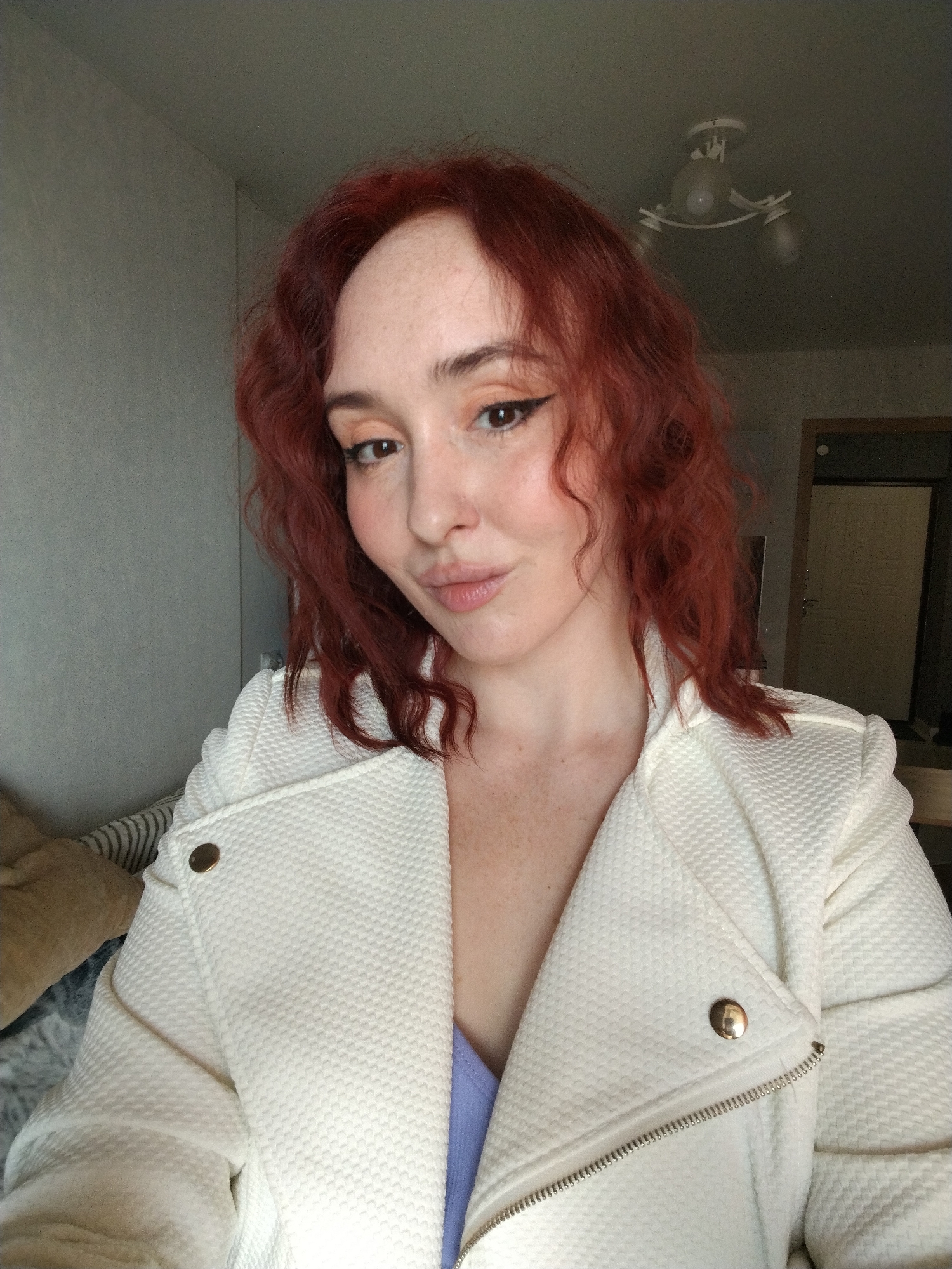 A woman with red curly hair and fair skin is posing for a selfie indoors. She is wearing a white textured jacket with gold buttons and a light purple top beneath. Her makeup includes winged eyeliner and peach-toned eyeshadow.
Behind her is a light gray wall and a partially visible doorway. A ceiling light fixture is visible in the upper right corner. There is a hint of bedding with a light brown pillow and striped pattern in the background to the left.
There is no visible information to suggest that this image was taken in Izhevsk, Russia. Therefore, the location of the image cannot be confirmed or denied to be in Izhevsk, Russia.
