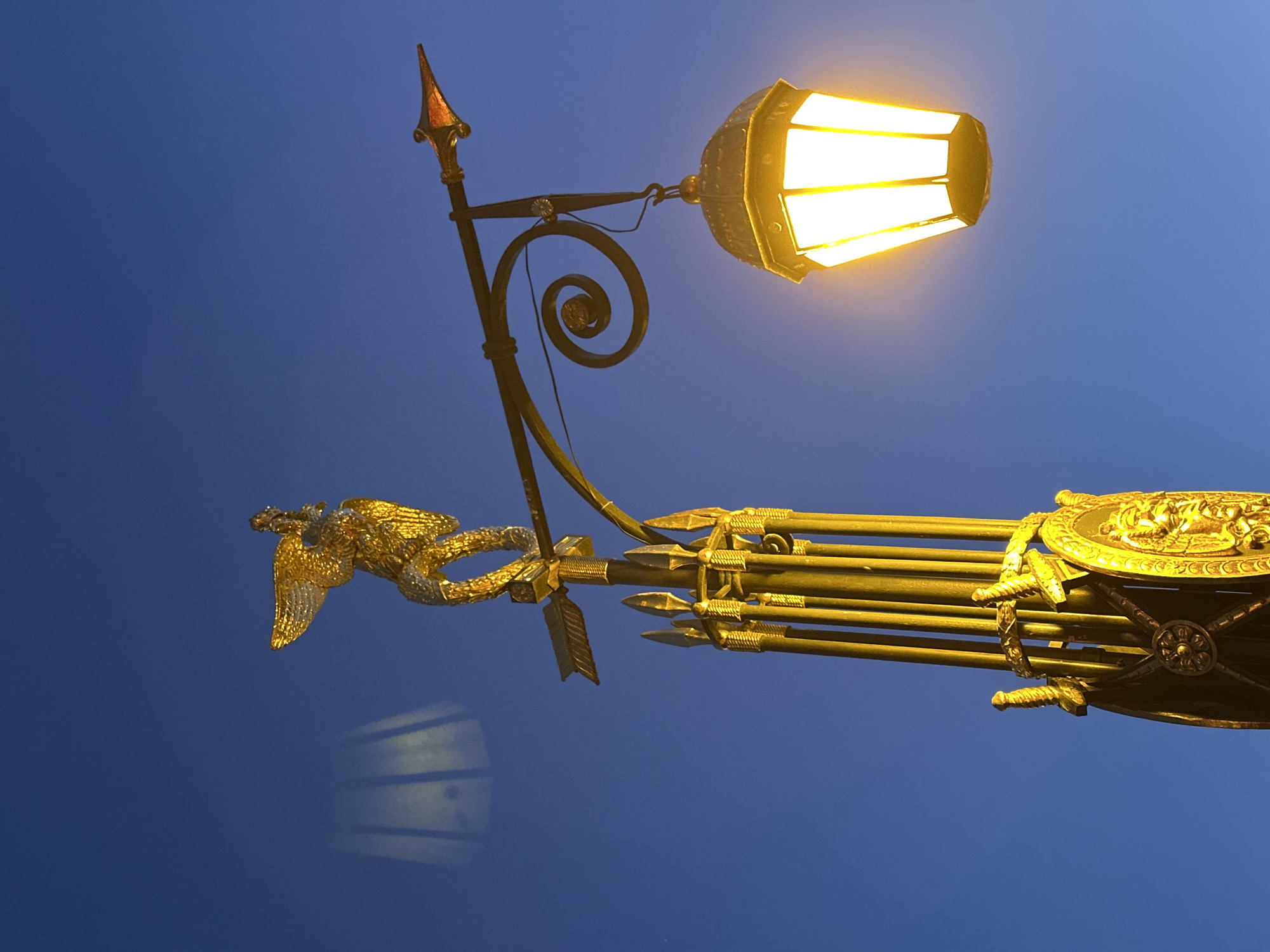 A low-angle view shows a golden decorative street lamp against a blue sky. The ornate lamp post features an eagle, arrows, a shield, and swords. The lamp itself is lit and casts a warm glow. This is a decorative feature of a bridge across the Neva River, St. Petersburg.