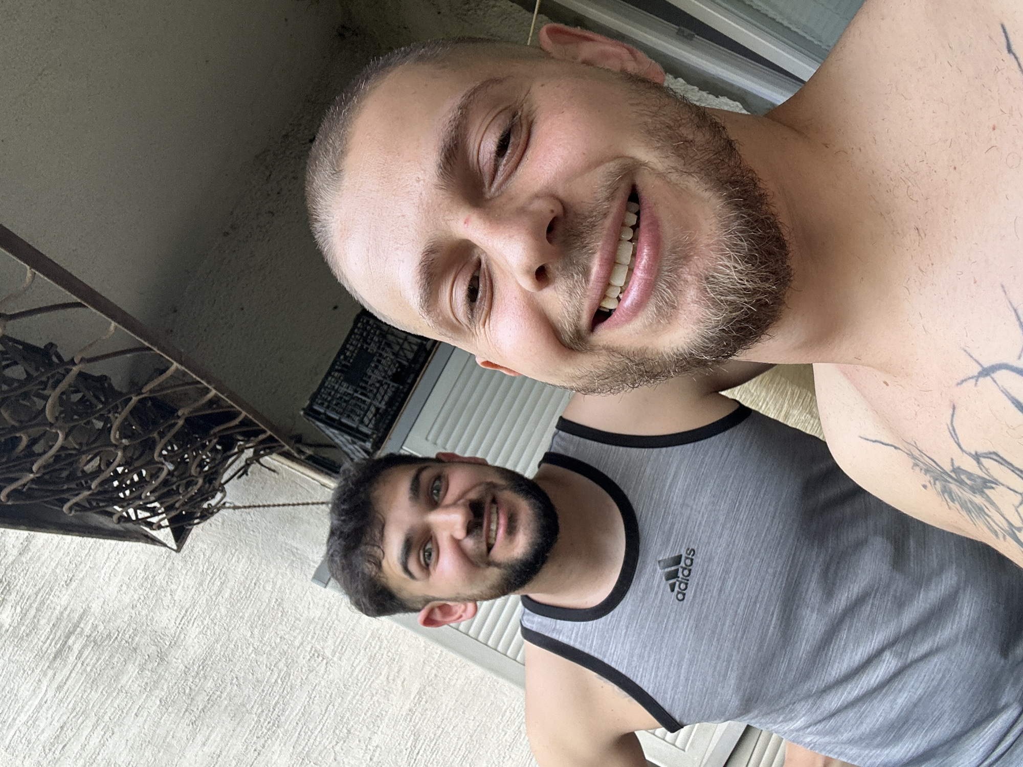 This media file is a selfie taken outdoors, featuring two young men. The man in the foreground is smiling widely and looking directly at the camera. He has short, buzzed hair, a beard, and a tattoo on his right arm. He is wearing a grey tank top with the Adidas logo on the left chest. The second man is partially visible in the background, also smiling. He has dark, curly hair and a beard.
The setting appears to be an outdoor area, possibly a balcony or patio, with a textured wall and some metal fencing visible. The lighting suggests it is daytime. The overall mood is casual and friendly, with both individuals appearing happy and relaxed. No specific event or activity is clearly depicted beyond the selfie itself. There is no discernible text other than the Adidas logo on the tank top.