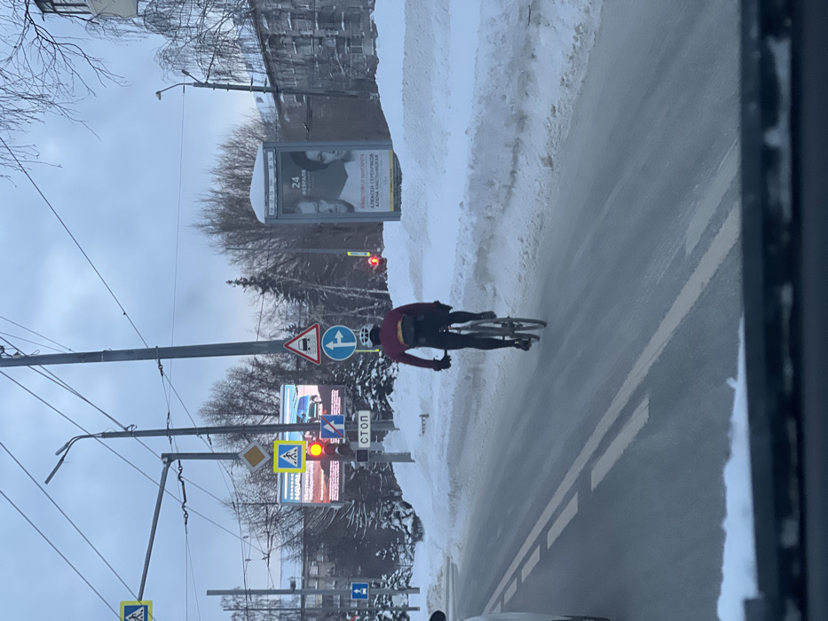 A cyclist is riding on a snowy street next to a snowbank. The sky is cloudy. There are road signs, a traffic light, and a billboard on the left side of the image. The traffic light is red. There are buildings and trees in the background. The image is taken from inside a vehicle, with a portion of the vehicle's interior visible on the right.