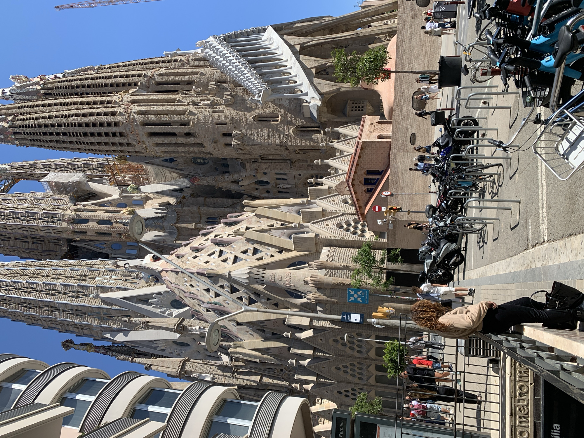 The image shows the Sagrada Familia basilica in Barcelona, Spain. The basilica is a large, unfinished architectural masterpiece with tall spires and intricate stone carvings. In the foreground, people walk along the sidewalk and there are parked bicycles. A person with curly hair rests on a metal railing near a "Metro" sign. The sky is clear and blue.