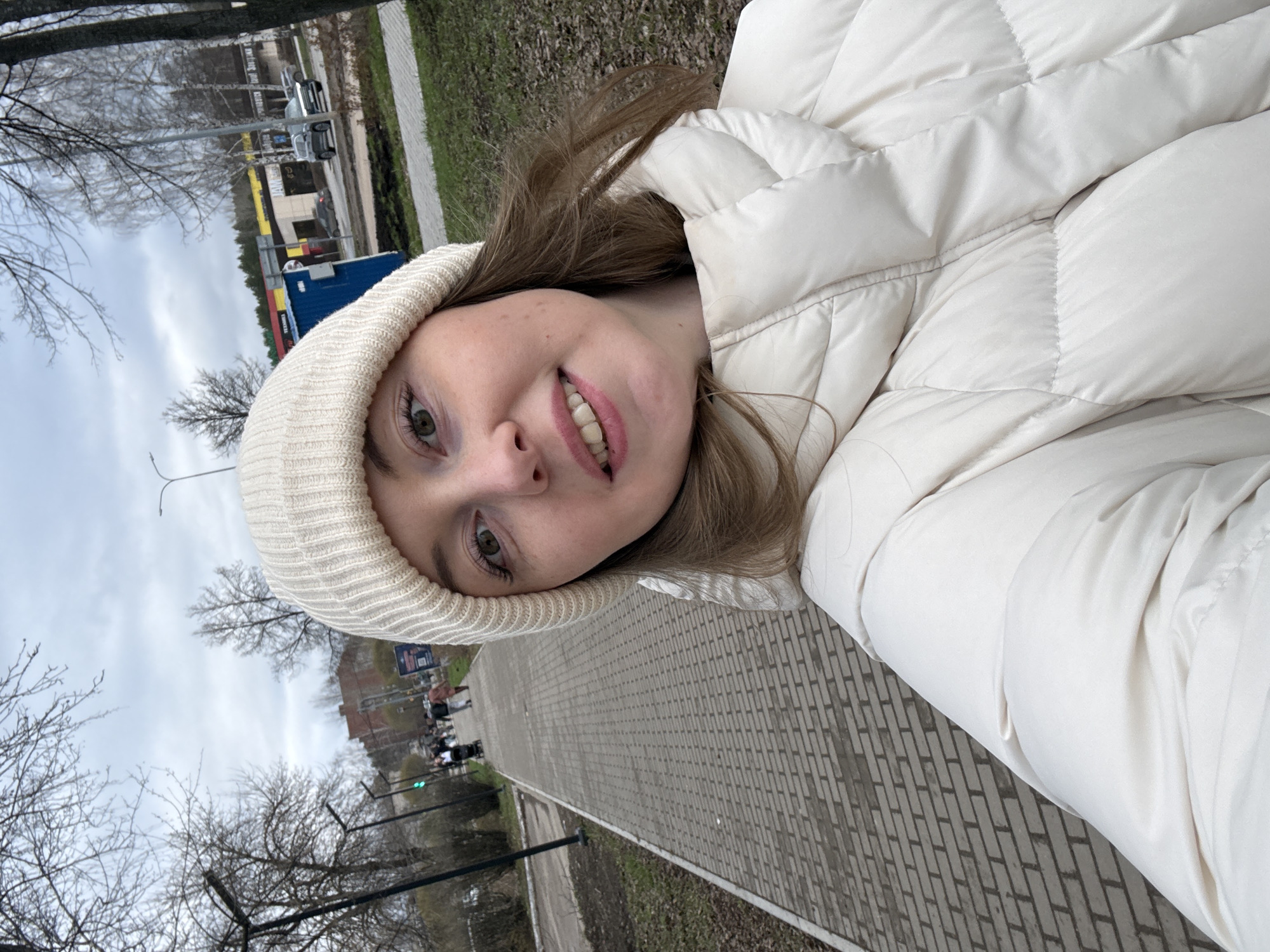 A young woman is featured prominently in the foreground of this selfie, angled slightly as she smiles towards the camera, revealing a subtle hint of teeth. She has light brown hair, green eyes, and light freckles across her nose and cheeks. She is dressed warmly in a light beige or off-white ribbed knit beanie and a matching puffer jacket, both suggesting cool weather.
The setting is an outdoor urban or suburban environment on an overcast day. A wide paved sidewalk, constructed from grey brick pavers, extends into the background on the right side of the image. To the left of the path, there are patches of grass and numerous bare trees, indicating either late autumn, winter, or early spring. In the mid-ground, a street runs parallel to the sidewalk, with a silver car and other vehicles visible. Further back, there are buildings, including one with distinctive yellow and blue sections, possibly a commercial structure. A lamppost and a green traffic light are also present in the background, along with blurry figures of other pedestrians. The sky is uniformly cloudy. No specific text is clearly legible in the scene.
