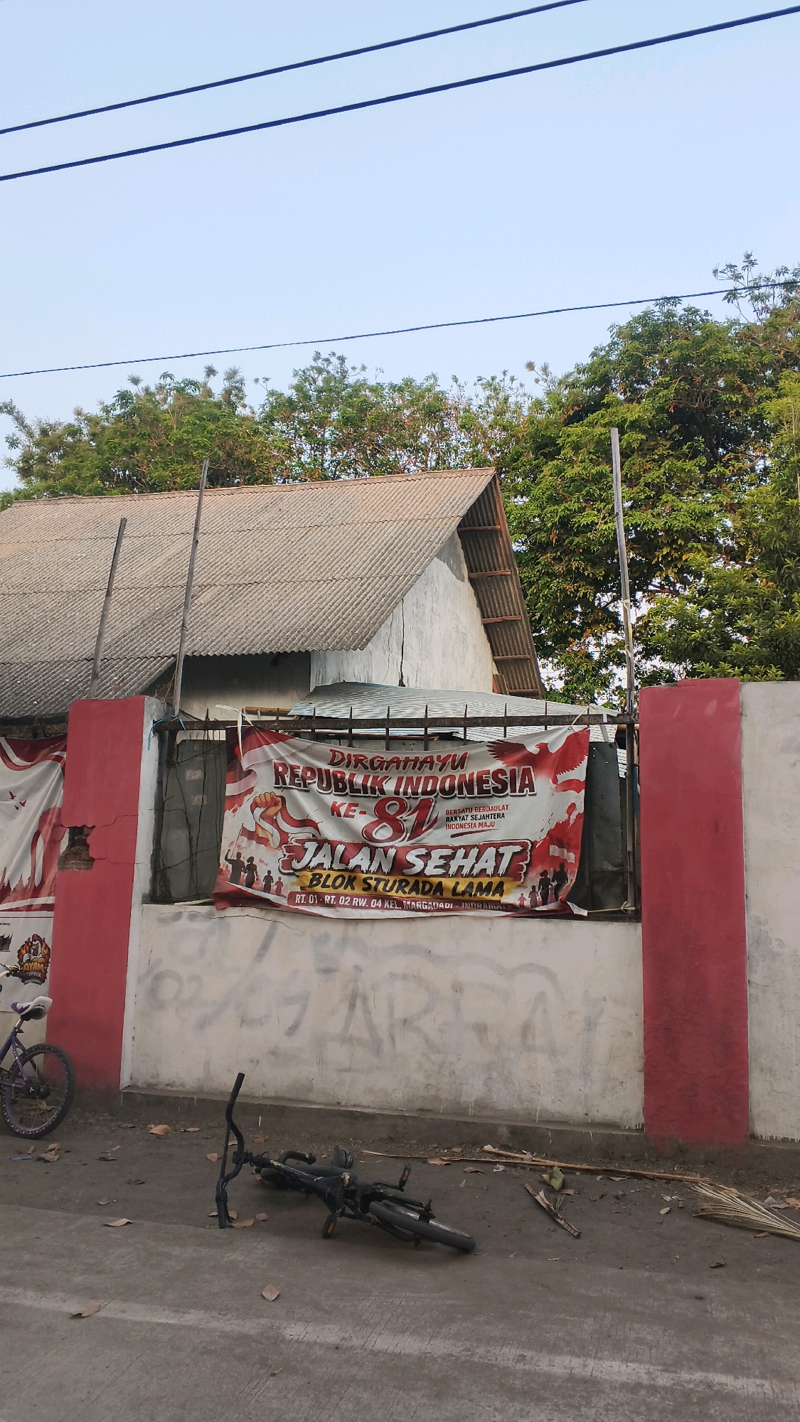 The image depicts a street scene in Indramayu, Indonesia. A large banner proclaiming "DIRGAHAYU REPUBLIK INDONESIA KE-81 JALAN SEHAT BLOK STURADA LAMA" is prominently displayed on a wall. Beneath the banner, graffiti is visible on the wall. In the foreground, a bicycle is lying on its side on the ground. Another bicycle is parked on the left side of the image. A house with a corrugated roof is visible behind the wall and banner. Trees are present in the background. The sky is clear and pale blue. The ground appears to be asphalt, with some scattered leaves and debris.