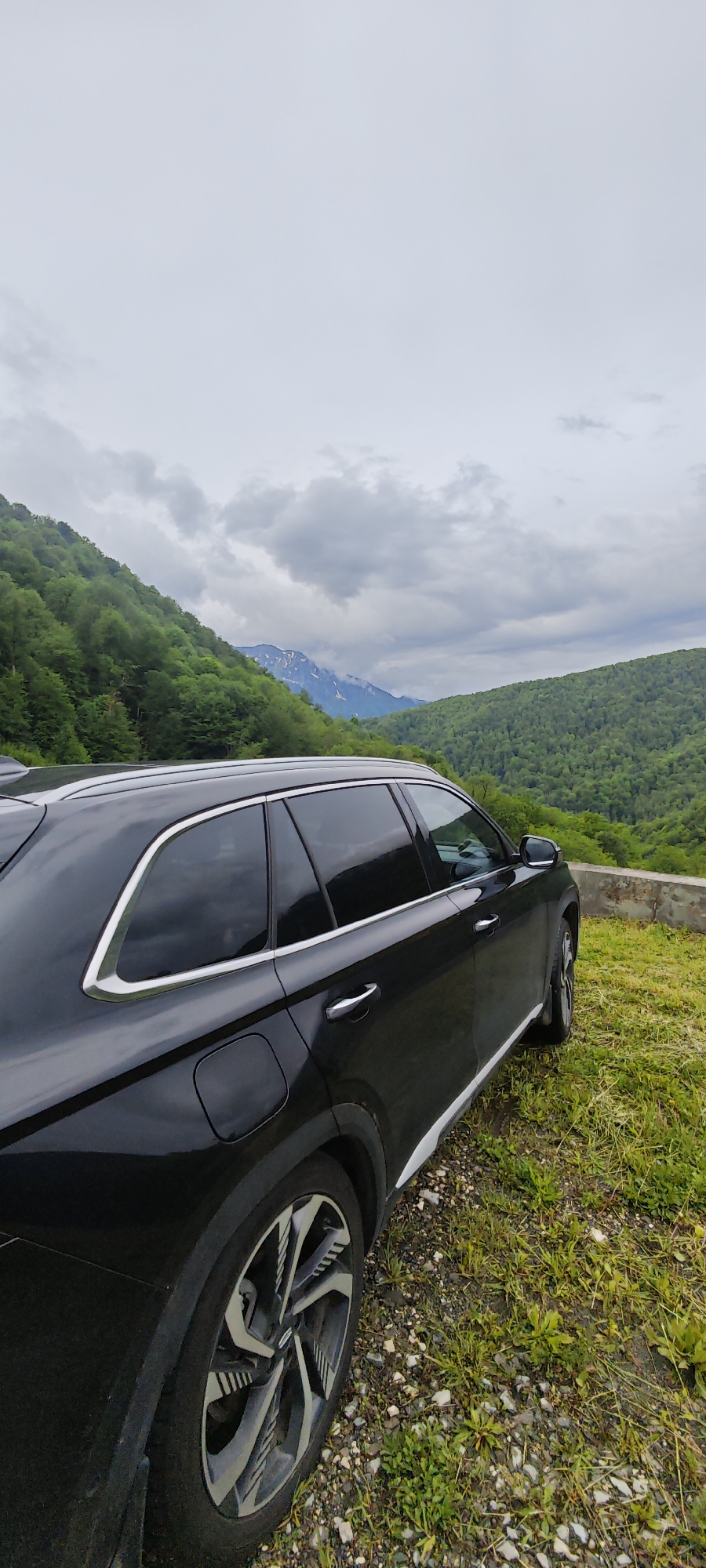 A black SUV stands parked on a grassy verge in Estosadok, Russia. In the background, forested hills rise towards a cloudy sky, with a distant mountain range visible. The focus is on the rear of the car, showing its tinted windows, silver trim, and a detailed wheel design. The ground beneath the car is a mix of grass, small stones, and patches of bare earth. A low concrete wall is partially visible behind the car on the right side. The atmosphere is overcast, suggesting a damp or cool day.