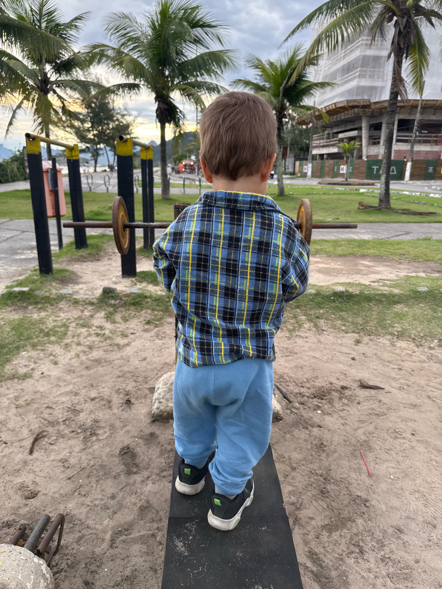 A young child stands facing away from the camera, on what appears to be an outdoor exercise area. The child is wearing a blue and black plaid shirt with yellow accents and light blue pants, and black sneakers with green accents. The child is standing on a black plank with a rough texture. In the background, there are palm trees, a construction site with a building covered in white scaffolding, and a cloudy sky. The ground is sandy with patches of grass. To the left, there is a weightlifting apparatus with yellow supports and a barbell with weights. This scene appears to be located in the city of Niterói, Brazil, possibly near a beach or park area.