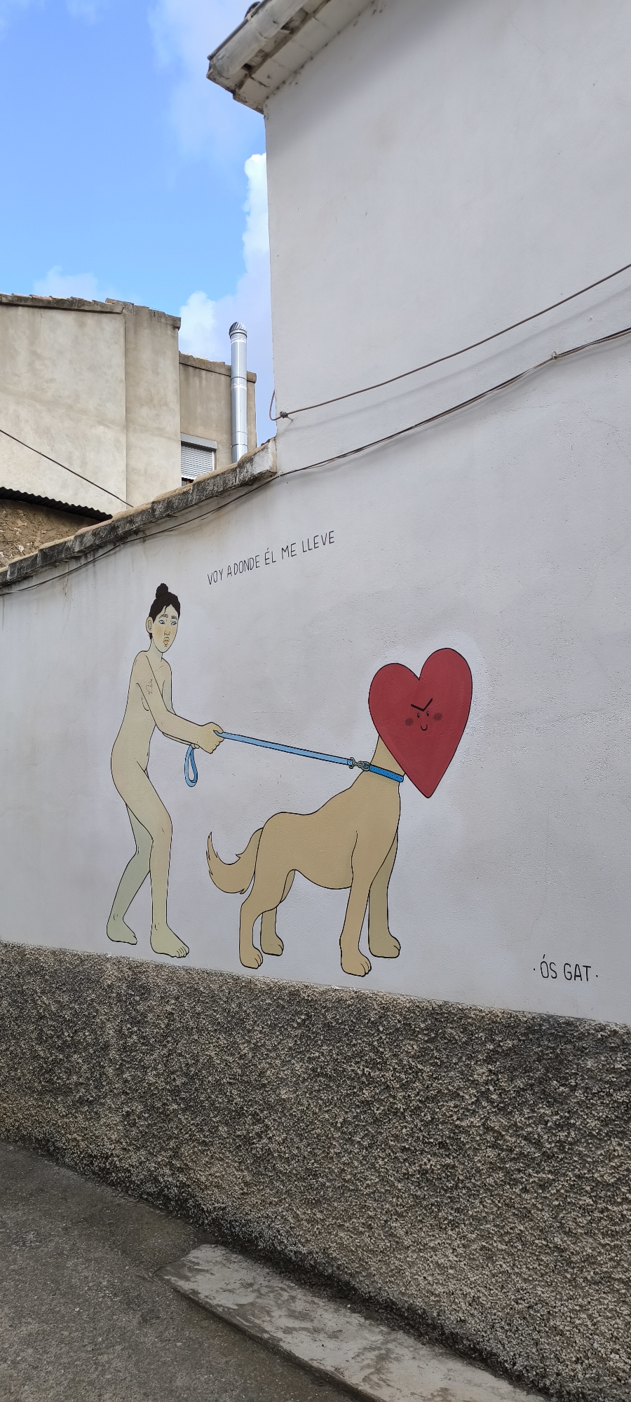 This image shows a mural on the exterior wall of a building in Fanzara, Spain. The mural depicts a stylized figure of a person walking a dog, where the dog's head is replaced by a red heart with a face. The figure is nude and is holding a blue leash attached to the heart-dog's collar. Above the mural, text in Spanish reads "VOY ADONDE ÉL ME LLEVE" which translates to "I go where he takes me." To the right of the mural, the signature "ÓS GAT" is visible. The wall is white, and a textured, gray base runs along the bottom of the mural. In the background, part of another building and the blue sky with white clouds are visible. The scene appears to be on a street or alleyway.