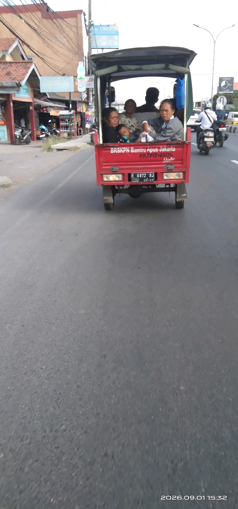 The image shows a red three-wheeled vehicle, likely a modified motorcycle, driving on a paved road in the city of Sunyaragi, Indonesia. The vehicle is carrying several passengers, including at least two adults and a baby. The adults appear to be elderly women, and the baby is being held by one of them, with a pacifier in its mouth. Another adult, a man, is visible in the background, possibly the driver. The vehicle has "BRSKPN Bambu Apus Jakarta" and "#KEMENSOS" printed on its side, suggesting it might be associated with a government or social welfare program. The road is lined with buildings and shops on the left side, with motorcycles parked in front of some of them. On the right side, other vehicles, including motorcycles, are also visible on the road. The overall scene depicts a common mode of transportation in Indonesia, with multiple people sharing a single vehicle.