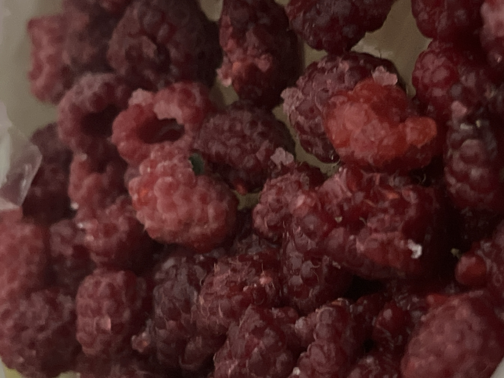 The image presents a close-up, top-down view of numerous raspberries, filling the majority of the frame. The raspberries exhibit a range of colors, from bright red to a deeper, darker purplish-red. Many of the berries are covered with small ice crystals, indicating they are either frozen or have recently been removed from a cold storage environment. The characteristic texture of raspberries, composed of individual drupelets, is clearly visible. Some of the berries are sharply focused, while others in the background are softly blurred, creating a shallow depth of field. A small, subtle green speck can be observed on one of the raspberries located towards the center-left. The background is out of focus and appears to be a light-colored surface, possibly the interior of a container or bowl. There are no people, discernible activities, or visible text in the image. The setting suggests an indoor environment, likely a kitchen or pantry.