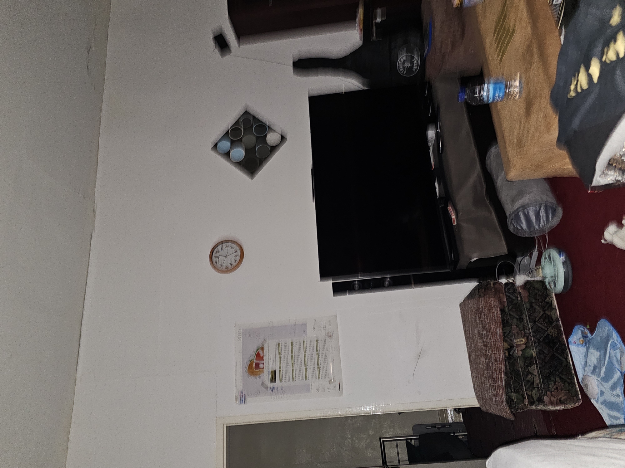 The image displays an indoor room, rotated 90 degrees clockwise, giving the impression that the walls are the floor and ceiling. The room features light-colored walls and a portion of a red carpet or rug on the floor.
Dominating the scene on one wall is a flat-screen television, positioned below a dark wooden furniture piece. Above the television, a dark wooden cabinet or shelf is also visible. On the adjacent wall, a round wooden-framed clock indicates the time is approximately 12:40. Below the clock, a calendar for the year 2016 is affixed, displaying months and dates. Above the calendar, a square recessed alcove contains multiple cylindrical objects in shades of blue, grey, and white, possibly decorative items or cups.
Numerous household objects are visible throughout the space. A blue plastic water bottle rests horizontally on a surface. Nearby, a brown paper bag, a grey cylindrical fabric bag, and a small, light blue fan or similar device are present. Towards the bottom of the frame, a patterned suitcase or storage box with brown and green floral designs is seen, alongside a partially visible white plush toy and a light blue fabric item. A white bedspread is also partly visible in the lower right corner. No people are depicted, and no specific activity is taking place. The room appears lived-in and somewhat cluttered, illuminated by what seems to be natural daylight. The location context is Holywell, United Kingdom.