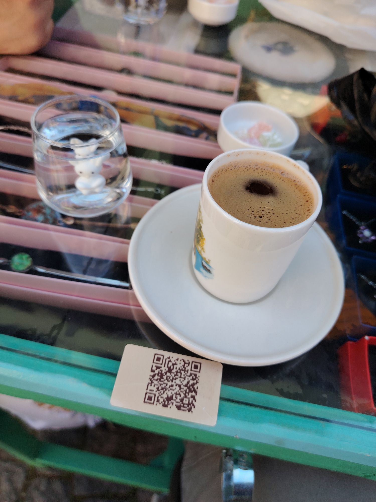 The image captures a close-up, eye-level view of a table setting, likely at a cafe or market stall in Eskisehir, Turkey. The main subjects are a small white ceramic cup of Turkish coffee, rich with dark foam, sitting on a matching saucer. Beside the coffee is a clear glass of water, which contains a small, white, stylized bunny figurine at its base.
In the blurred background, several other items are visible. To the left and behind the coffee cup, there is a small white bowl holding colorful, pastel-hued sweets, resembling Turkish delight. Further back, various pieces of jewelry, including necklaces with different stones (one green, one purple), are displayed on pink-lined trays and in blue compartments. A person's arm is slightly visible in the top-left corner, suggesting a customer or server. Part of a white dish with a blue design and a black bag are also discernible in the background.
The setting appears to be outdoors or semi-outdoors, possibly a sidewalk cafe or a stall in a marketplace, given the casual arrangement and the display of goods. The table itself is made of glass with a green frame underneath. A small, rectangular card bearing a QR code is placed on the table in the lower-left foreground, likely for payment or information. The natural lighting suggests it is daytime. The overall scene depicts a moment of relaxation, enjoying traditional Turkish coffee with its customary accompaniment of water and sweets, amidst a commercial or cultural environment.