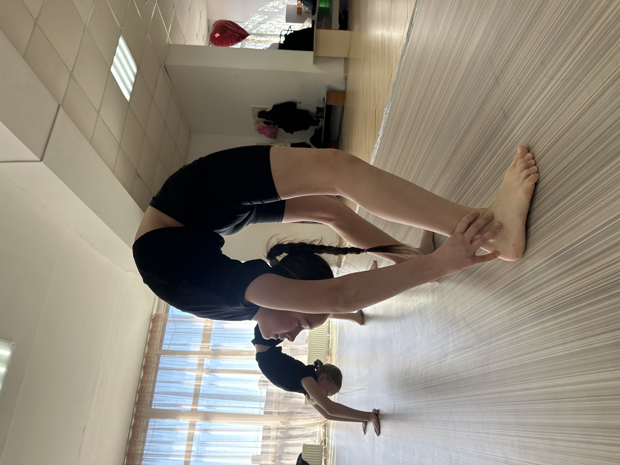 This image captures two individuals, likely young females, practicing gymnastic backbends or "bridges" within an indoor facility in Votkinsk, Russia. The foreground subject, wearing a black t-shirt and black shorts with dark hair styled in two braided pigtails, is performing a deep backbend, reaching her hands back to hold her lower legs. A second, smaller individual in black is visible further back, also executing a backbend with hands on the floor.
The setting appears to be a bright studio or gym, characterized by a light-colored wooden floor, white walls, and a tiled ceiling with fluorescent lighting. Large windows adorned with sheer curtains line one wall, allowing natural daylight to illuminate the space, suggesting it is daytime with clear weather outside. A red heart-shaped balloon is partially visible in the upper background, near a window. The scene conveys a focused training environment. There is no visible text in the image.