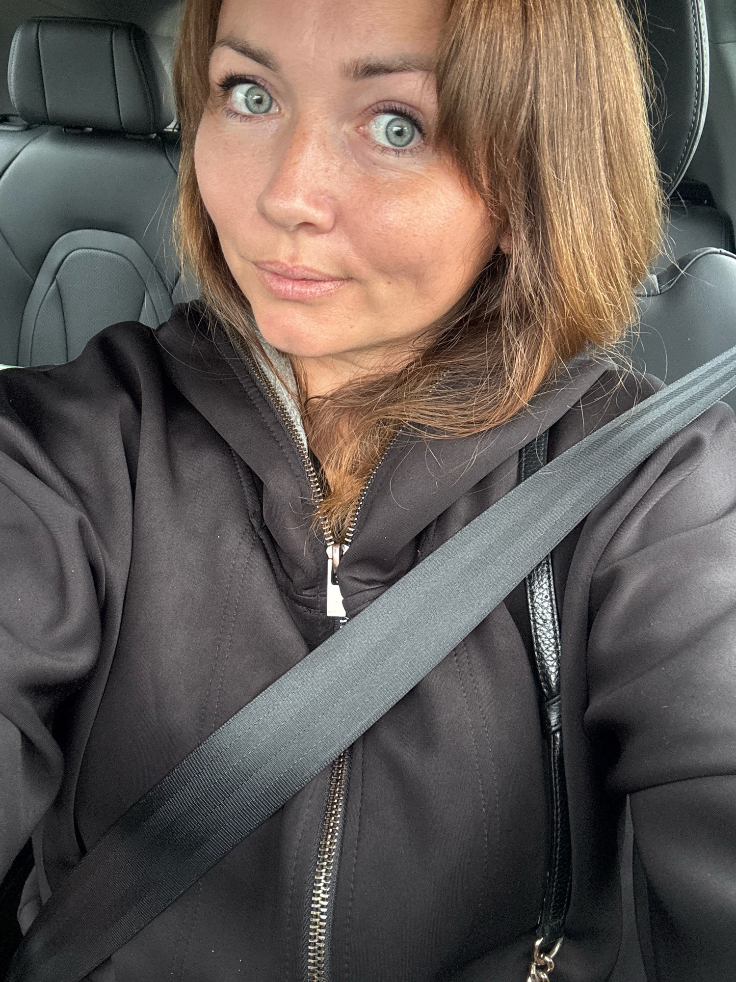 The image shows a woman sitting in the driver's seat of a car. She is wearing a black hooded jacket over a gray shirt and has a seatbelt across her chest. The interior of the car appears to be dark leather. The woman has light skin with freckles, blue eyes, and brown hair. She is looking at the camera with a slight smile. The background consists of the car's headrest and interior. The image does not contain any information about the city of Timmolovo, Russia.