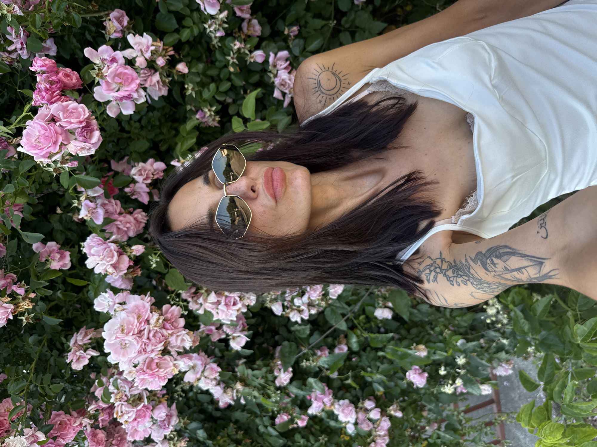 A woman with dark hair, wearing sunglasses and a white dress, poses for a selfie in front of a bush with pink flowers. She has tattoos on her arm and shoulder. The image was taken in the city of Pleven, Bulgaria.