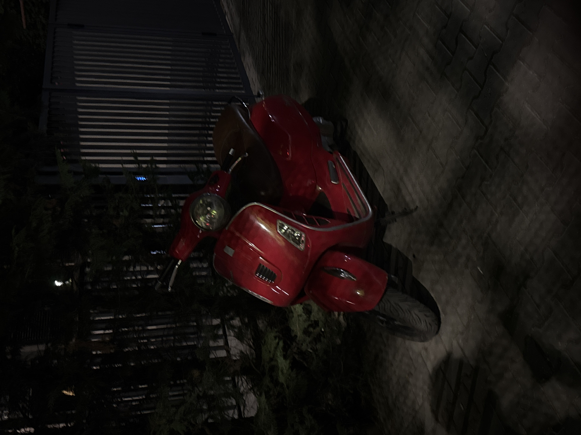 A red Vespa-style scooter is parked on a patterned paver sidewalk at night. The scooter is facing left, with its front wheel turned slightly. A dark green coniferous shrub is behind the scooter, and to the right, there is a dark gray metal gate with vertical slats. The lighting suggests an outdoor, possibly residential or commercial, area in Sofia, Bulgaria. No people are present in the image.