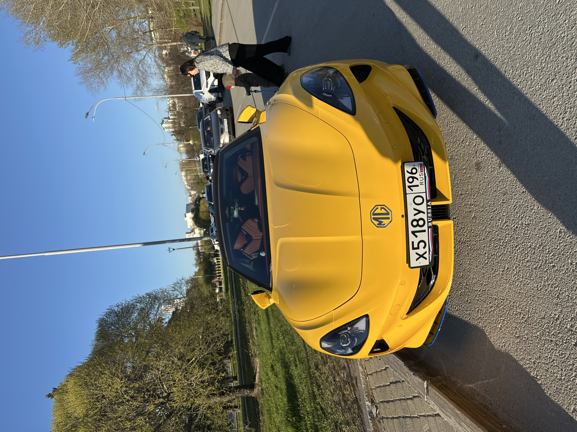 The image shows a yellow MG TF LE500 convertible parked on a paved road adjacent to a grassy area with trees. The car has a Russian license plate "X518YO 196 RUS." A person in a patterned jacket and dark pants is standing near the car, seemingly interacting with another person who is partially visible. In the background, there is a line of parked cars and a lamppost under a clear blue sky. The scene appears to be taken during daylight. It is very likely that this scene is in Yekaterinburg, Russia based on the license plate.