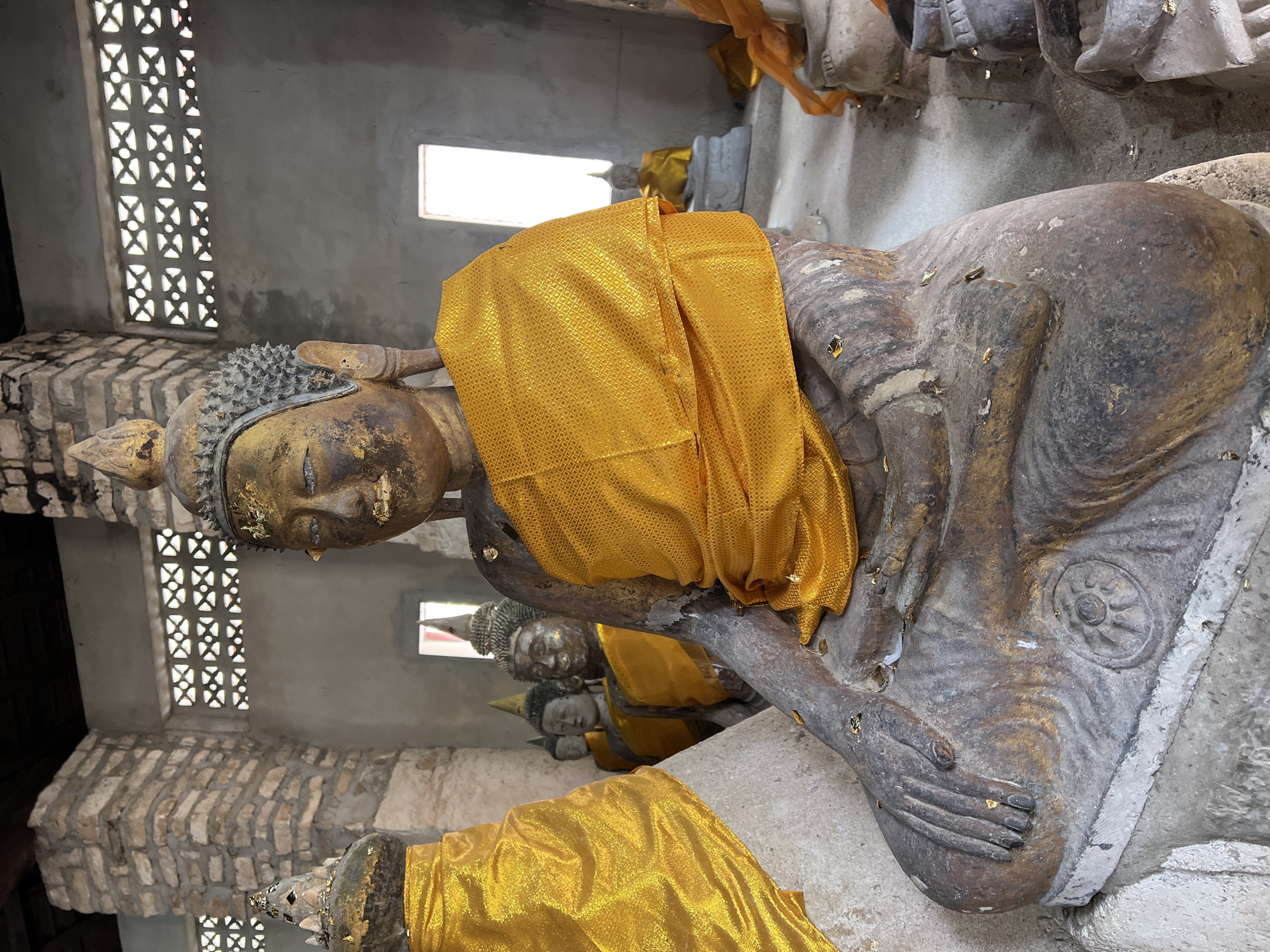 In Baan Harn, Thailand, within a weathered structure with stone and concrete walls and a dark wooden ceiling, a weathered, seated stone Buddha statue is prominently featured in the foreground. The statue wears a vibrant yellow cloth draped over its shoulder. Its face, weathered and tinged with gold, looks forward, with closed eyes and a serene expression. Small flecks of gold leaf adorn the statue's skin and fabric. Behind this central figure, several other similar Buddha statues are visible, also draped in yellow cloth, receding into the background. The environment appears to be a temple or shrine, with windows or openings allowing diffused light into the space. The floor is a rough concrete surface, and the overall impression is one of age and reverence.