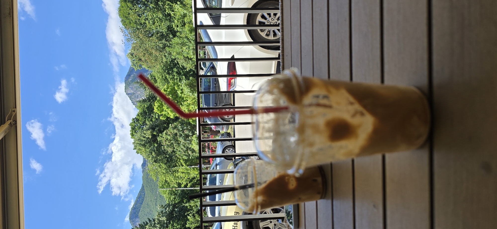 A bright, clear day unfolds in this image, with a vibrant blue sky dotted with fluffy white clouds. The foreground is dominated by a wooden table, upon which sit two partially consumed iced drinks in clear plastic cups, each with a straw. One cup, in the foreground, is out of focus and shows a heart-shaped mark on its side. The other cup, slightly further back and in focus, reveals a dark straw.
Beyond the table, a black metal railing separates the viewing area from a street. Parked cars are visible behind the railing, including a white car with a distinct silver rim, a dark gray SUV, and a silver sedan. A yellow vehicle with the partial word "Happy" visible on its side is also present.
The middle ground is filled with lush green trees, their leaves appearing healthy and verdant. Behind the trees, majestic mountains rise, their peaks etched against the sky. The overall impression is one of a peaceful outdoor setting, perhaps a cafe patio, offering a scenic view of nature.