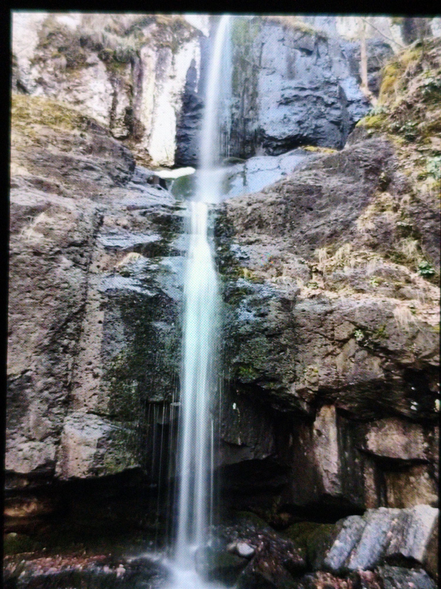 The image depicts a waterfall in Devin, Bulgaria. Water cascades down a rocky cliff face covered in moss. The rocks are various shades of grey and brown. The waterfall appears as a blurred white streak due to long exposure. The foreground is dark and shadowed, with some rocks visible at the base of the falls.