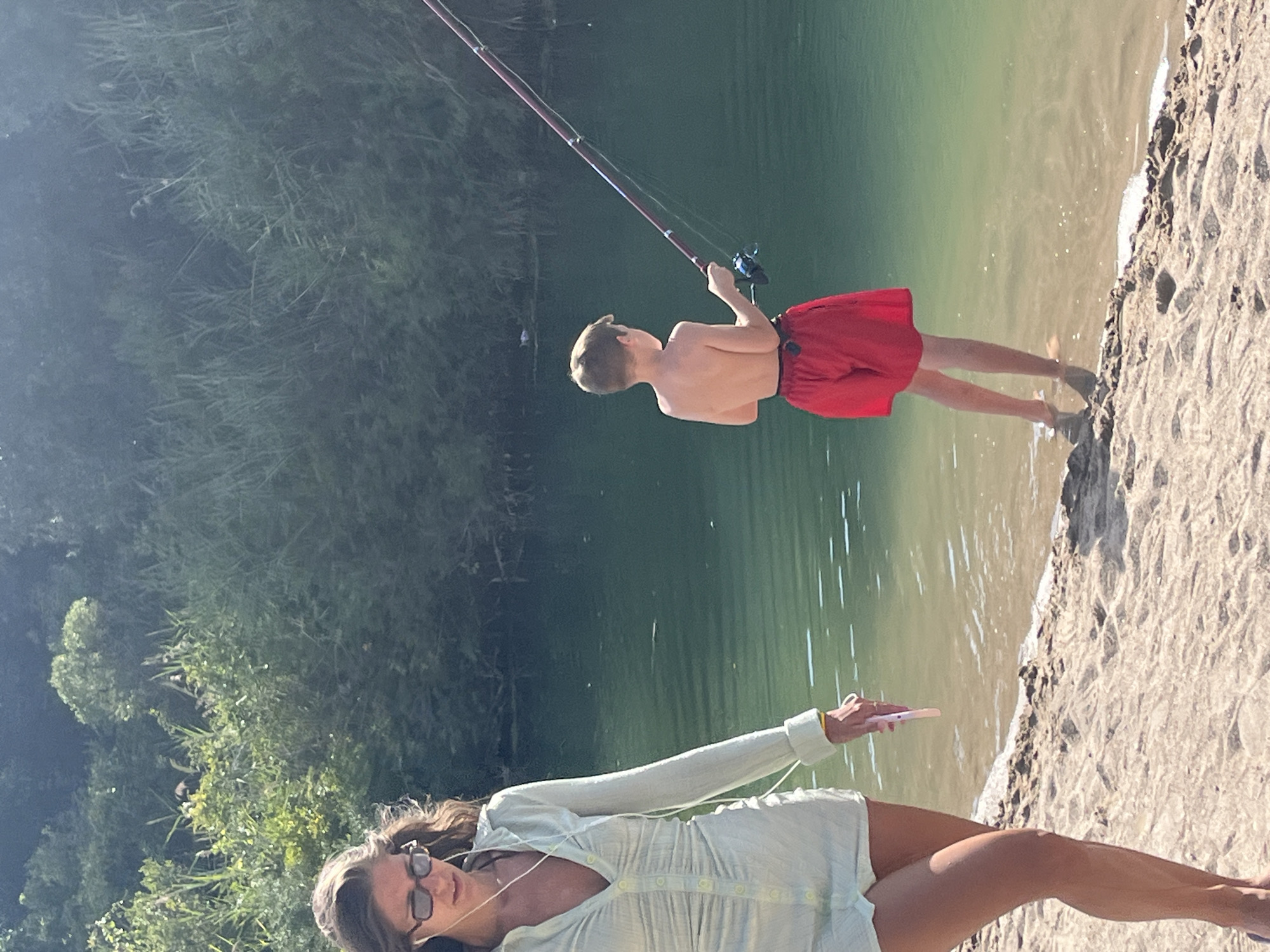 The image shows a young boy fishing in a body of water, possibly a river or lake, near a sandy shore. He is wearing red shorts and is holding a fishing rod. To the left of the boy, a woman is walking along the shore. She has on a light-colored dress and is wearing sunglasses and earphones. Behind them, lush green vegetation lines the opposite bank of the water. The lighting suggests it is daytime, with sunlight illuminating the scene. The specific location in Prijevor, Montenegro is not identifiable from the image itself, but the setting appears to be a natural outdoor environment with water and plant life.
