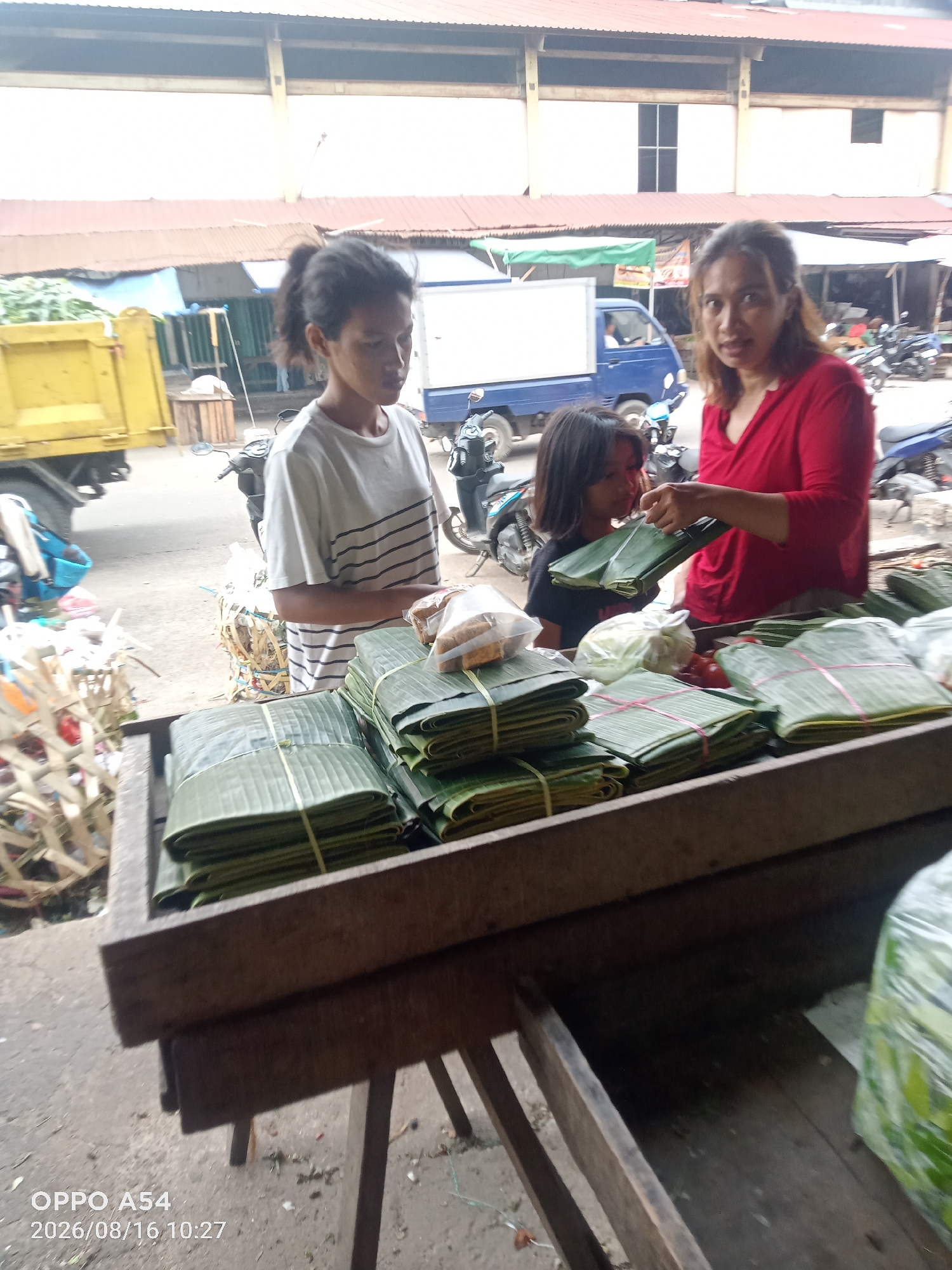 In Jambi City, Indonesia, three individuals are present at an outdoor market stall. A woman in a red shirt is standing, holding a bundle wrapped in green leaves, interacting with a young girl. The young girl, wearing a black t-shirt, is looking at the item in the woman's hand. Another young woman with her hair in a ponytail, wearing a striped t-shirt, stands to the left of the stall, facing away from the camera. The stall is laden with numerous packages wrapped in green leaves, secured with string. In the background, a yellow truck is visible, along with market structures and parked motorcycles. The scene suggests a transaction or display of goods at a local market.