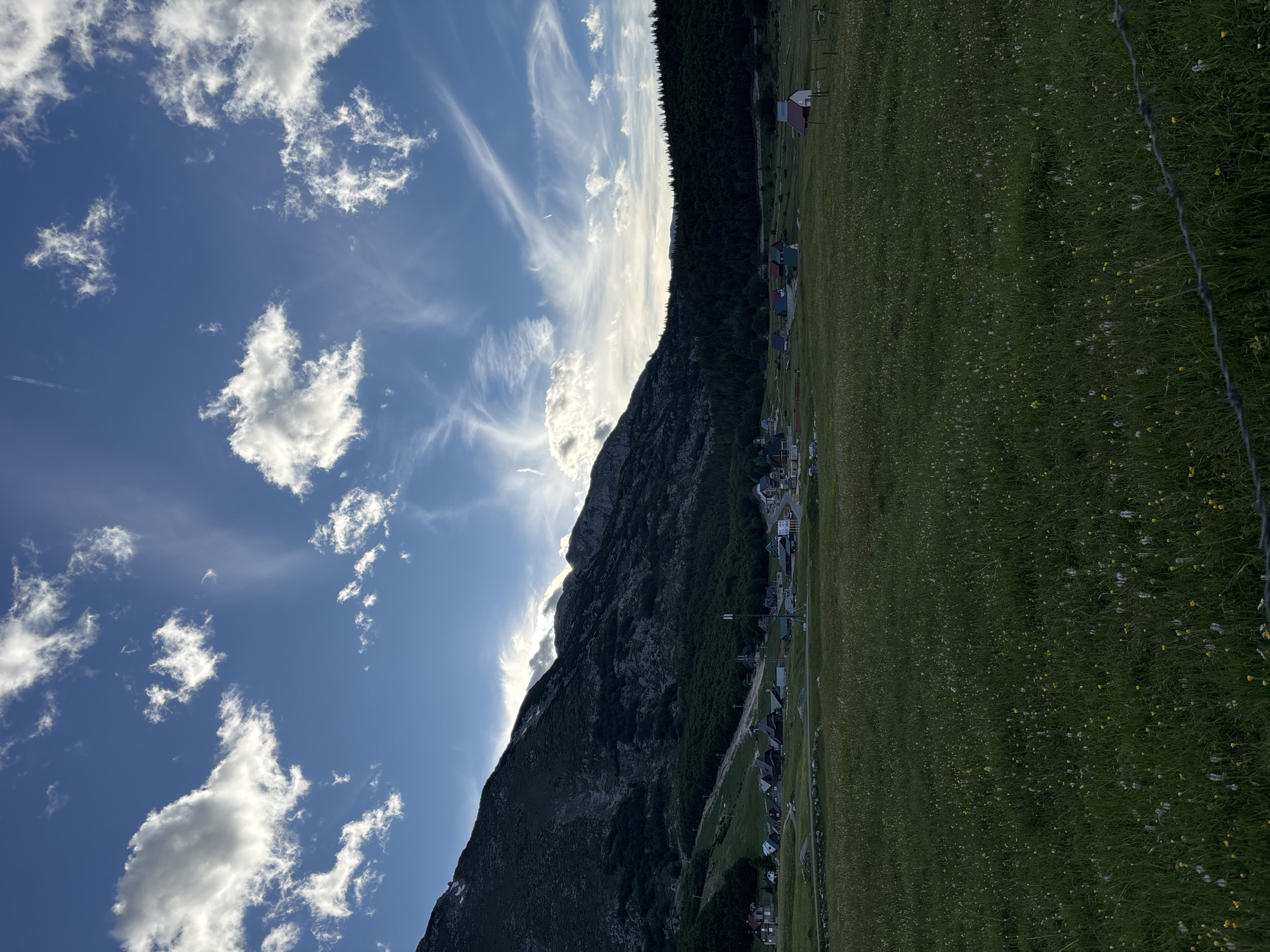 This is an outdoor, vertical, eye-level shot of a meadow in front of a mountain range near the village of Virak, Montenegro. The sky is blue with scattered white clouds. The foreground is a grassy field with small yellow and white flowers. A barbed wire fence is visible in the bottom right corner. In the middle ground, there is a cluster of small houses and buildings. The background features a dark, forested mountain range. The overall lighting suggests it is either late afternoon or early morning. There are no people or animals visible in the image.