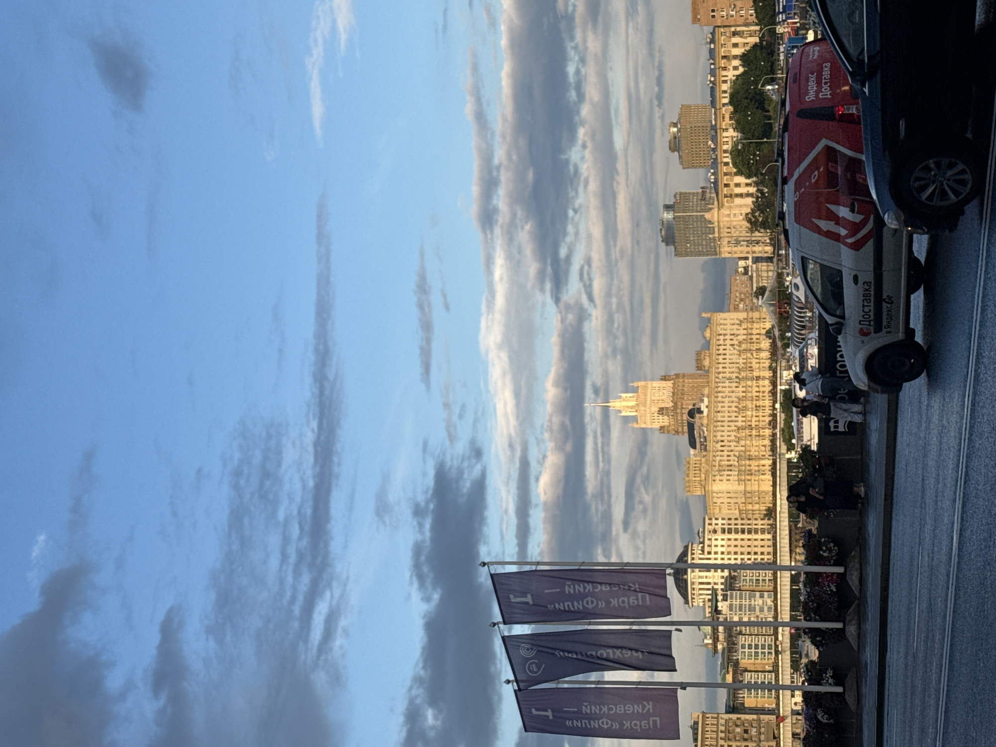 In Moscow, Russia, on a cloudy day, a view of the city skyline is presented. In the foreground, a wet road reflects the ambient light, with two vehicles in motion. A white van with red branding and the text "Яндекс Доставка" (Yandex Delivery) is visible, alongside a black car. Behind the vehicles, a row of three purple flags with white lettering hangs from poles. Further in the background, several buildings of varying architectural styles form the city's panorama, including a prominent, ornate yellow building topped with a spire. Several people are standing on a walkway in front of these buildings, some appearing to be engaged in conversation. The sky above is a blend of blue with scattered white and gray clouds.