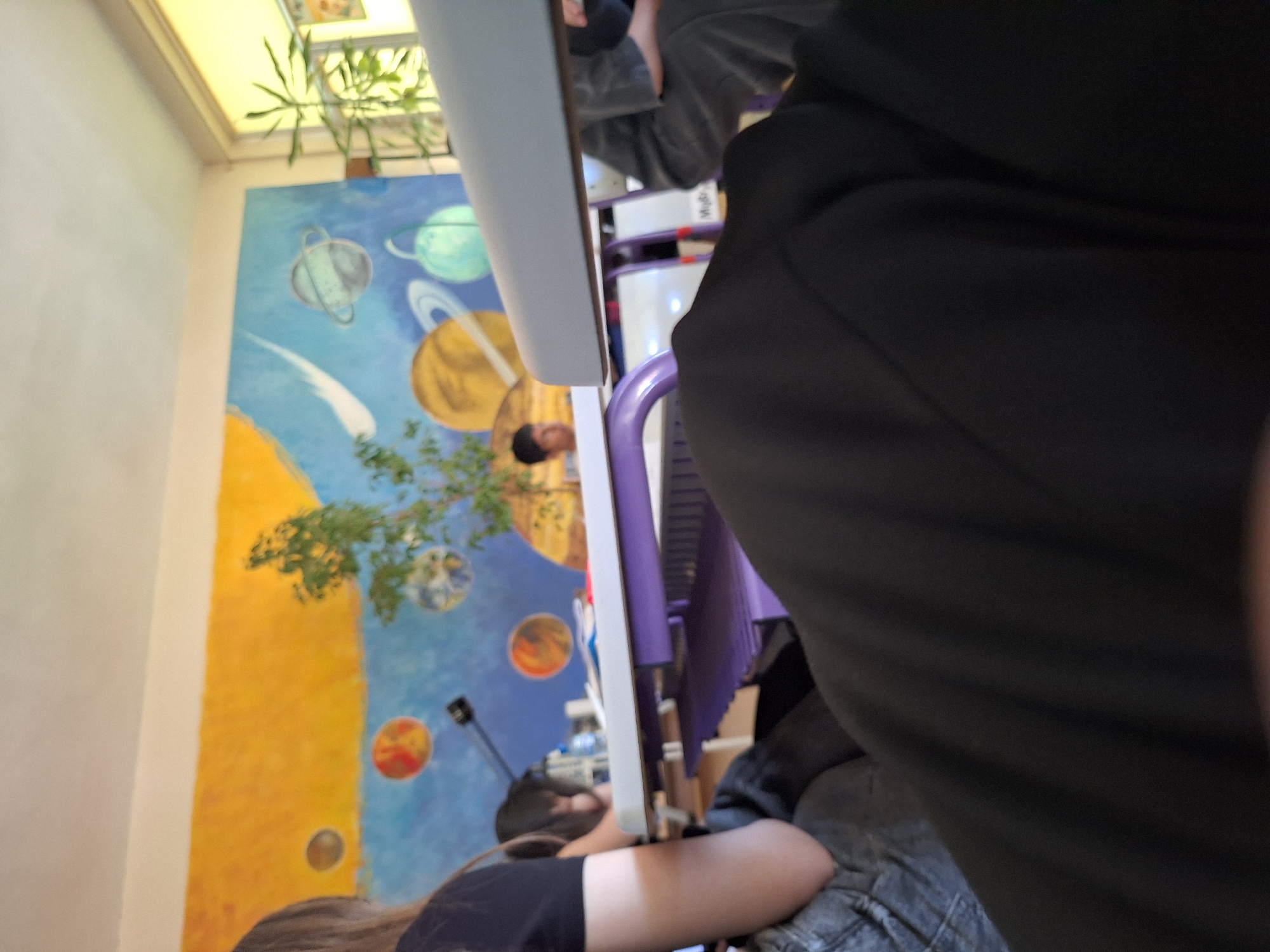 The image depicts the interior of a classroom, likely at a school, featuring a mural on the back wall with a space theme, including planets and a yellow sky. In the foreground, a person's legs covered in black fabric are prominently visible, suggesting a first-person perspective. A desk with purple legs is in the middle ground, with papers and a water bottle on its surface. Behind the desk, another person, a young male with dark hair, is partially visible. To the left, a person with long blonde hair wearing a black t-shirt is also visible, facing away from the camera and towards the desk. A plant is situated to the right of the frame, near a window. The lighting suggests it is daytime. The location is stated to be in Varna, Bulgaria.