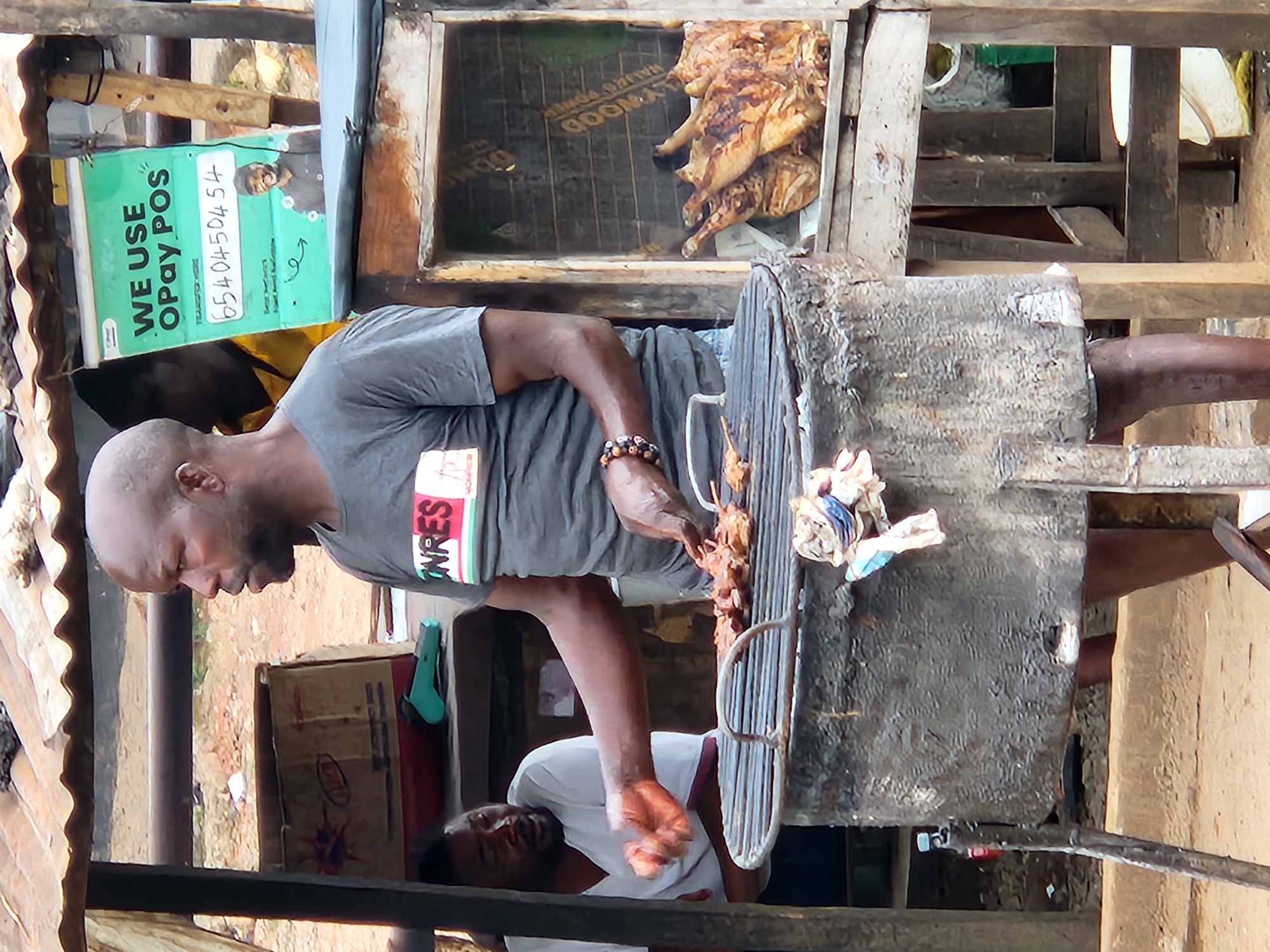 A mid-shot shows a bald Black man wearing a grey t-shirt with a red and white logo that says "ONRES". He is standing and appears to be cooking meat on a charcoal grill. His right arm is raised, and his hand is over the grill, holding tongs. He is looking to the right. Behind him, another Black man in a white t-shirt is visible, looking at the first man. To the right of the first man, a wooden structure houses a display of cooked chickens. A green sign in the background reads "WE USE Opay POS" and displays a phone number. The scene appears to be an outdoor market stall, likely in Jos, Nigeria, given the context of "Opay POS" which is a common payment method in the region. The ground is dirt, and there is a corrugated metal roof overhead, suggesting a makeshift stall. There are also various boxes and other items scattered around the stall. The overall impression is one of a bustling local marketplace.