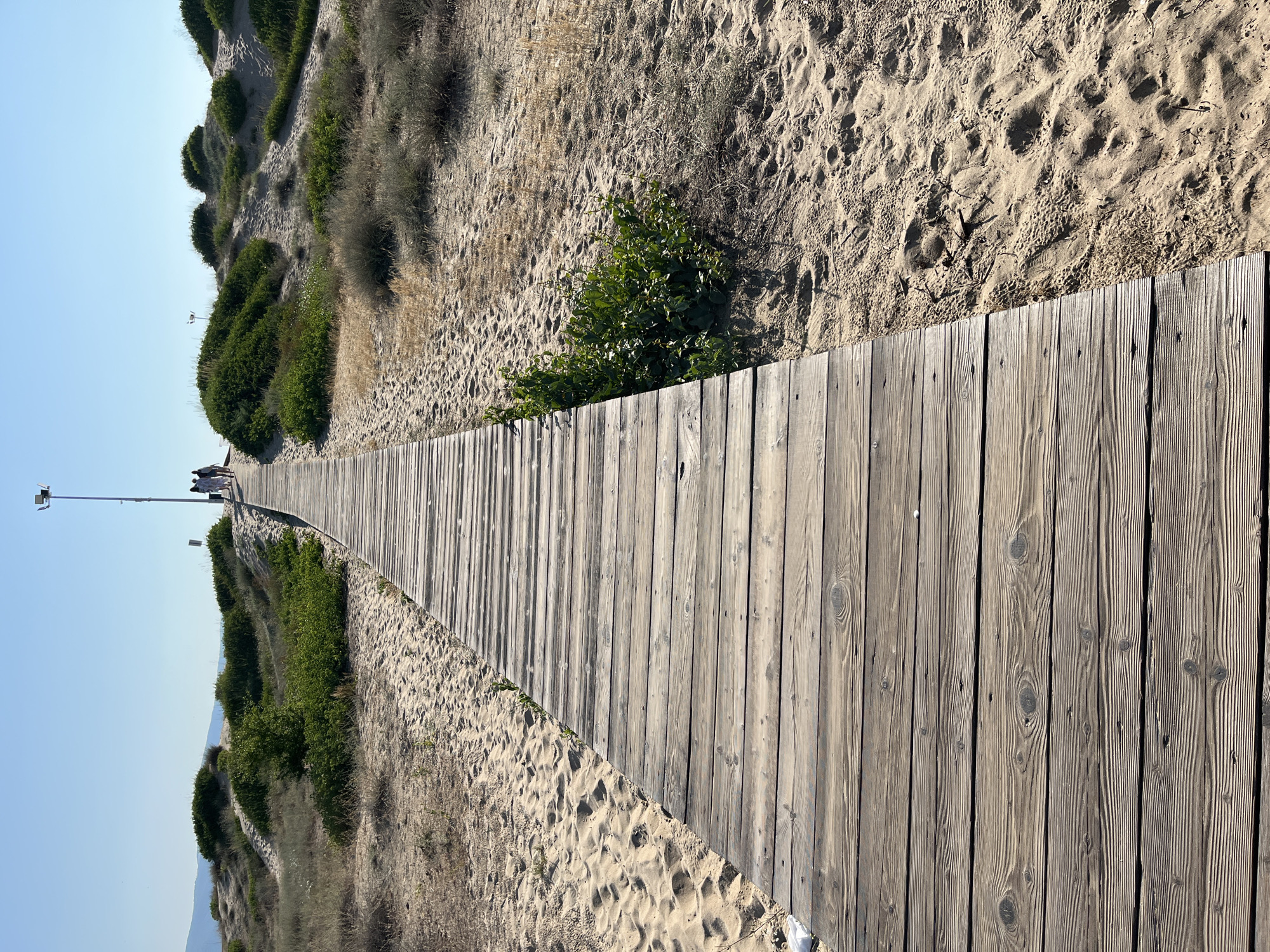 A wooden boardwalk extends into the distance across a sandy dune landscape. The boardwalk is made of weathered wooden planks running horizontally, with sand visible on either side. Low green shrubbery and dry grasses are scattered across the dunes. In the far distance, a lamppost stands on the right side of the boardwalk, and a few people are walking away from the camera along the path. The sky is clear and blue, indicating a sunny day. The scene is likely a coastal area, and specifically the area around the resort of Sunny Beach, near Nesebar, Bulgaria.