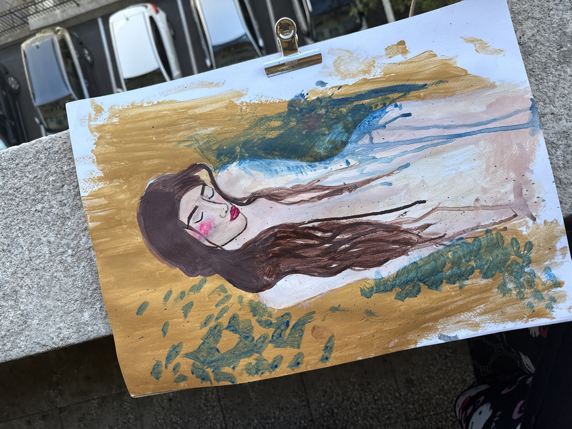A horizontal image captures a drawing of a woman lying on her side. The woman has dark hair, fair skin, and pink cheeks. The artwork is positioned outside on what appears to be a ledge or low wall. The background includes several cars parked along the street. The exact location in the city is unknown.