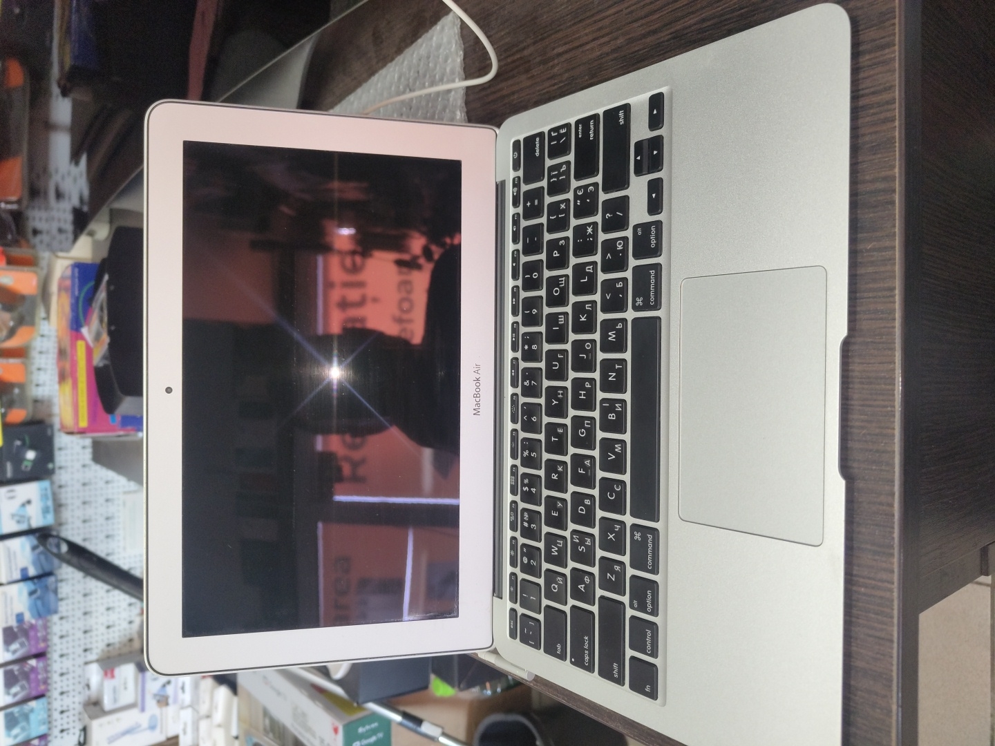 This image shows a close-up, rotated view of an open MacBook Air laptop, resting on a dark wooden surface. The laptop is silver with a black keyboard and a large trackpad. The screen displays a reflection of an indoor environment, with blurred shapes and colors suggesting shelves with products. Text within the reflection appears to be in a non-Latin script and also includes English words such as "area", "Retie", and "efoa". The "MacBook Air" text is visible on the bottom bezel of the laptop. The keyboard layout appears to be for a Cyrillic alphabet, commonly used in Moldova, where Chișinău is the capital. There are no people visible in the image. The lighting suggests an indoor setting, possibly a store or a home, and there are no strong indicators of the time of day or weather. The overall scene depicts a solitary object in a cluttered background, with the focus on the device itself.