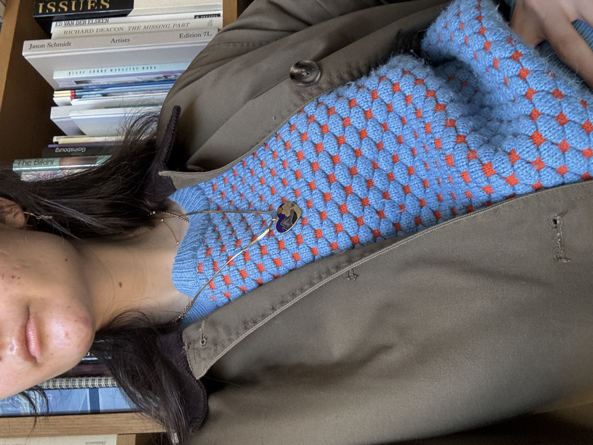 The image depicts a person lying down, with their cheek resting on a stack of books. The book titles visible on the spines include "ISSUES," "ED VAN DER ELSKEN," "RICHARD DEACON THE MISSING PART," "Jason Schmidt Artists Edition 7L," "The Bikini," and "Earth Sk."
The person is wearing a blue sweater with a patterned texture and orange diamond-shaped designs. A gold necklace with a small charm is visible around their neck. They are also wearing a green jacket.
The setting appears to be indoors, possibly a library or home, based on the presence of the bookshelves and books. There is no direct indication that the location is in New York City.