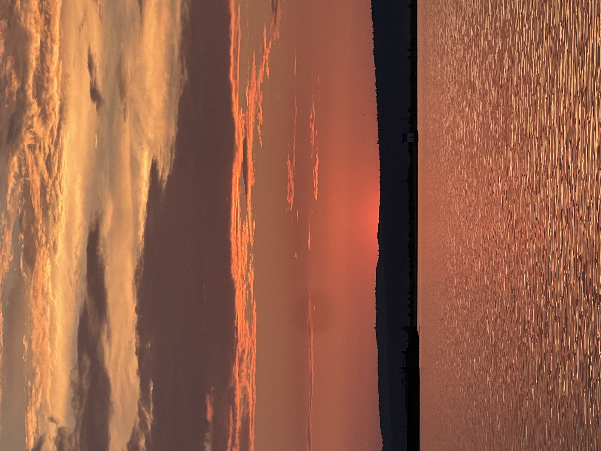The image captures a sunset over the Kama River in Chaikovsky, Russia. The sky is a vibrant blend of orange, red, and purple hues, with scattered clouds reflecting the fading sunlight. A dark, silhouetted line of hills forms the horizon, with a few indistinct structures visible along its edge. The calm surface of the river mirrors the fiery colors of the sky, creating a picturesque and serene scene.