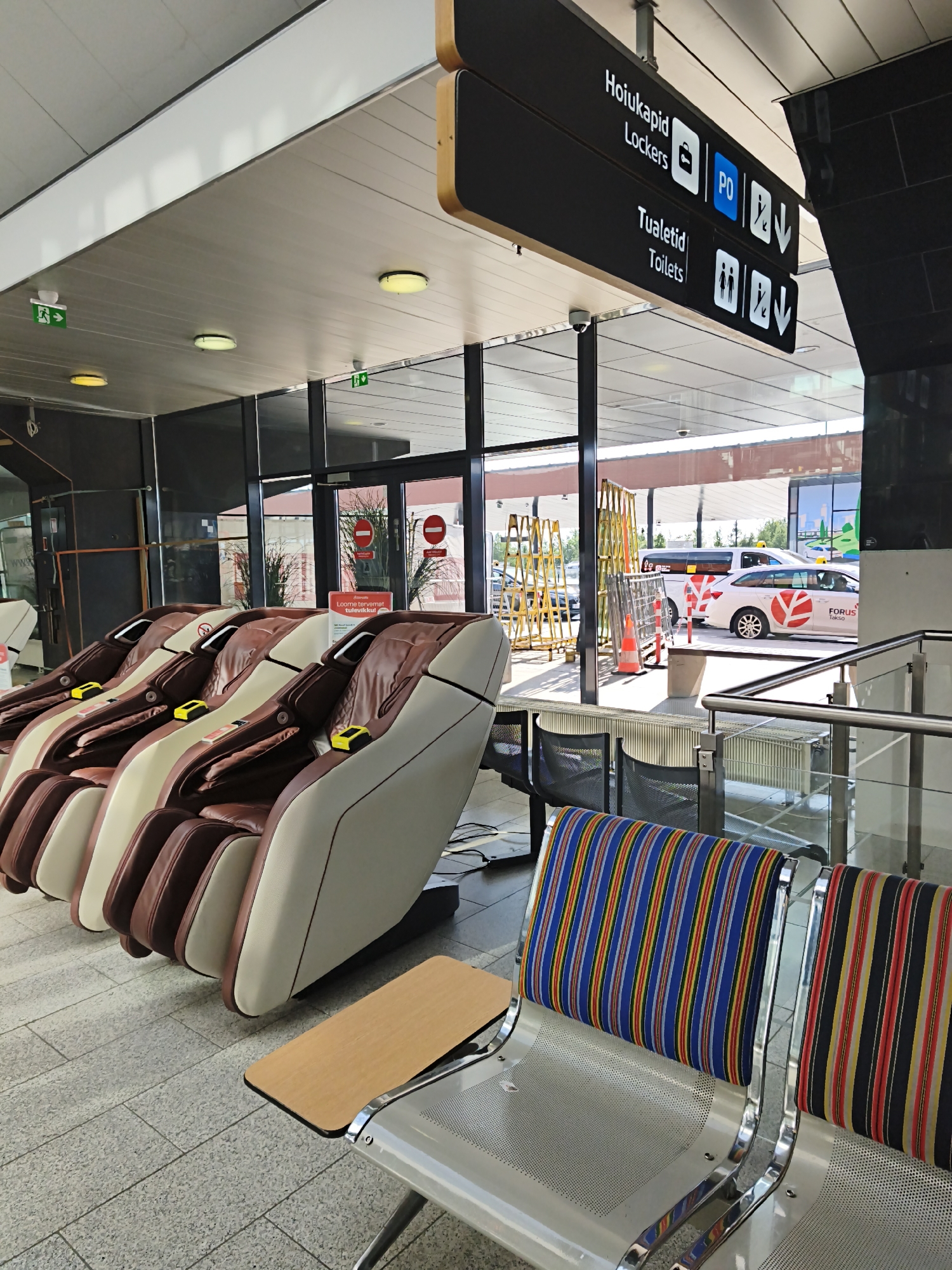 The image shows a waiting area with several massage chairs and a row of metal benches with striped cushions. There is a sign overhead indicating "Hoiukapid Lockers" and "Tualetid Toilets." In the background, through a glass wall, there is a taxi and some traffic. The floor is tiled. The scene appears to be inside a public building, possibly an airport or transportation hub, in Tallinn, Estonia. There are no people visible in the image.