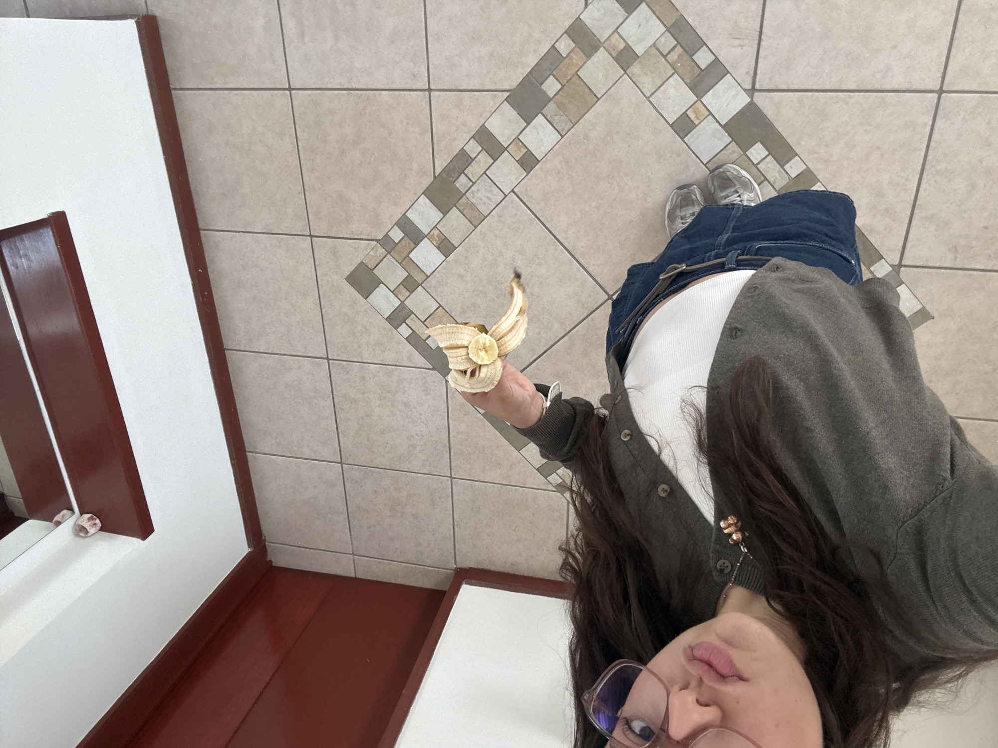 The image, captured from a high-angle perspective looking downwards, features a young woman standing indoors on a tiled floor. Her head is tilted, and she gazes upward towards the camera with a neutral expression, her lips slightly parted. She has long dark hair and wears clear-framed glasses, a white ribbed top, a dark gray button-up cardigan, and blue jeans cinched with a dark belt. Her feet are clad in silver-colored sneakers.
In her right hand, she holds a partially peeled banana, with one round slice still attached to the peel. A gold-toned necklace with a clustered pendant is visible around her neck.
The floor is composed of light beige square tiles, interrupted by a decorative diagonal band of smaller, alternating light and dark gray and beige square tiles. To her left, there is a white structure with dark reddish-brown wooden trim, which includes a mirror. A small, light-colored, possibly shell-like object rests on the edge of this mirror. The setting appears to be a well-maintained indoor space, potentially in Heroica Puebla de Zaragoza, Mexico, under bright, even daytime lighting. No visible text is present in the image.