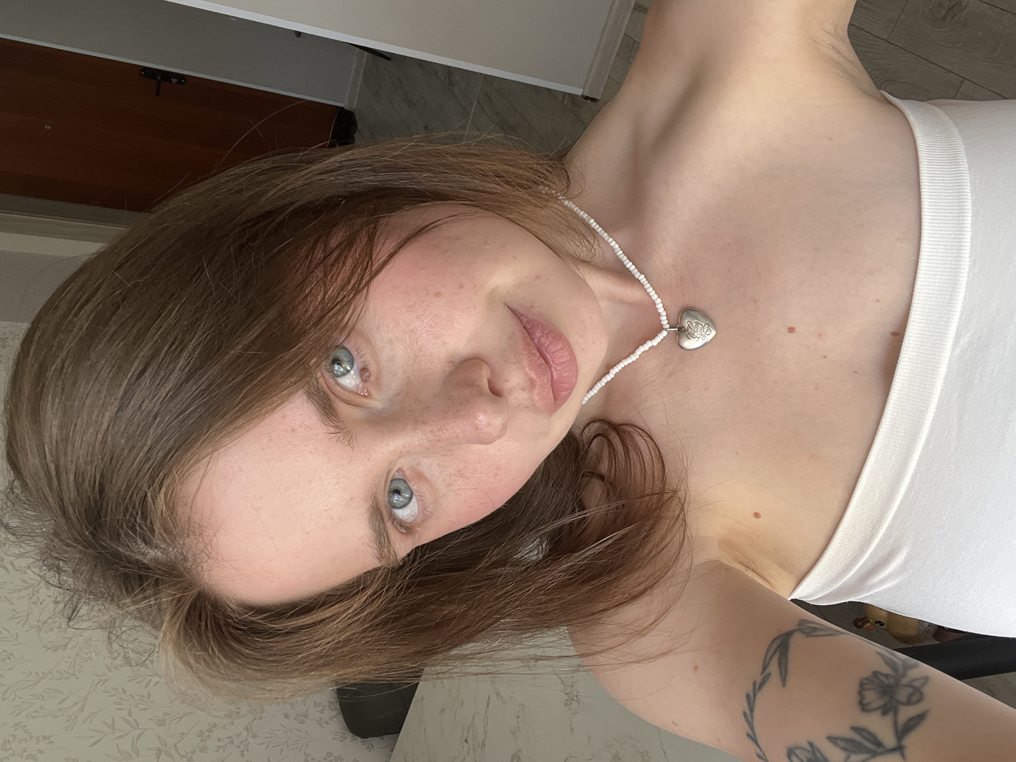 A young Caucasian woman with light brown hair and blue eyes looks directly at the camera. She is wearing a white strapless top and a beaded necklace with a heart-shaped pendant. A tattoo of floral branches is visible on her left arm. The background appears to be an indoor setting with a patterned wallpaper, a door, and flooring. The lighting suggests it is daytime.