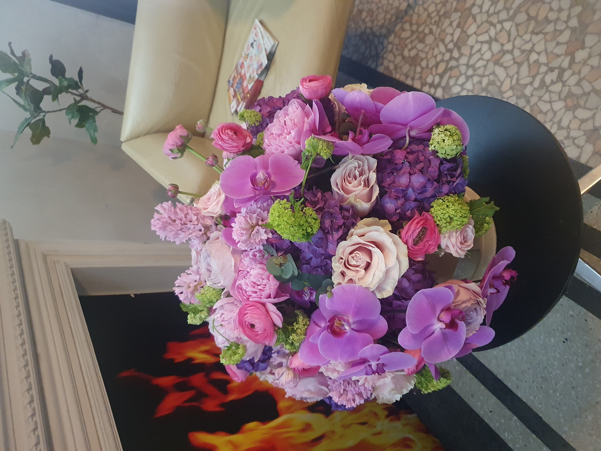 A large, elaborate floral arrangement occupies the center foreground, resting on a round, black table with a metallic base. The bouquet is densely packed with various flowers in shades of pink, purple, lavender, and green, including prominent Phalaenopsis orchids, hydrangeas, ranunculus, roses, hyacinths, and small clusters of bright green florets. To the left background, a white, ornate fireplace mantelpiece frames an active artificial fire display exhibiting orange and yellow flames against a dark background. The floor in front of the fireplace features dark tiles with two prominent light-colored horizontal stripes. To the right background, a cream-colored upholstered sofa or armchair is partially visible, with a stack of colorful magazines or brochures placed on its cushion. Behind the sofa, a tall, slender potted plant with green leaves is positioned against a light-colored wall. The floor to the right consists of light-toned terrazzo or mosaic tiles. No individuals are present. The specific location within Plovdiv, Bulgaria, is not discernible from the provided visual evidence.