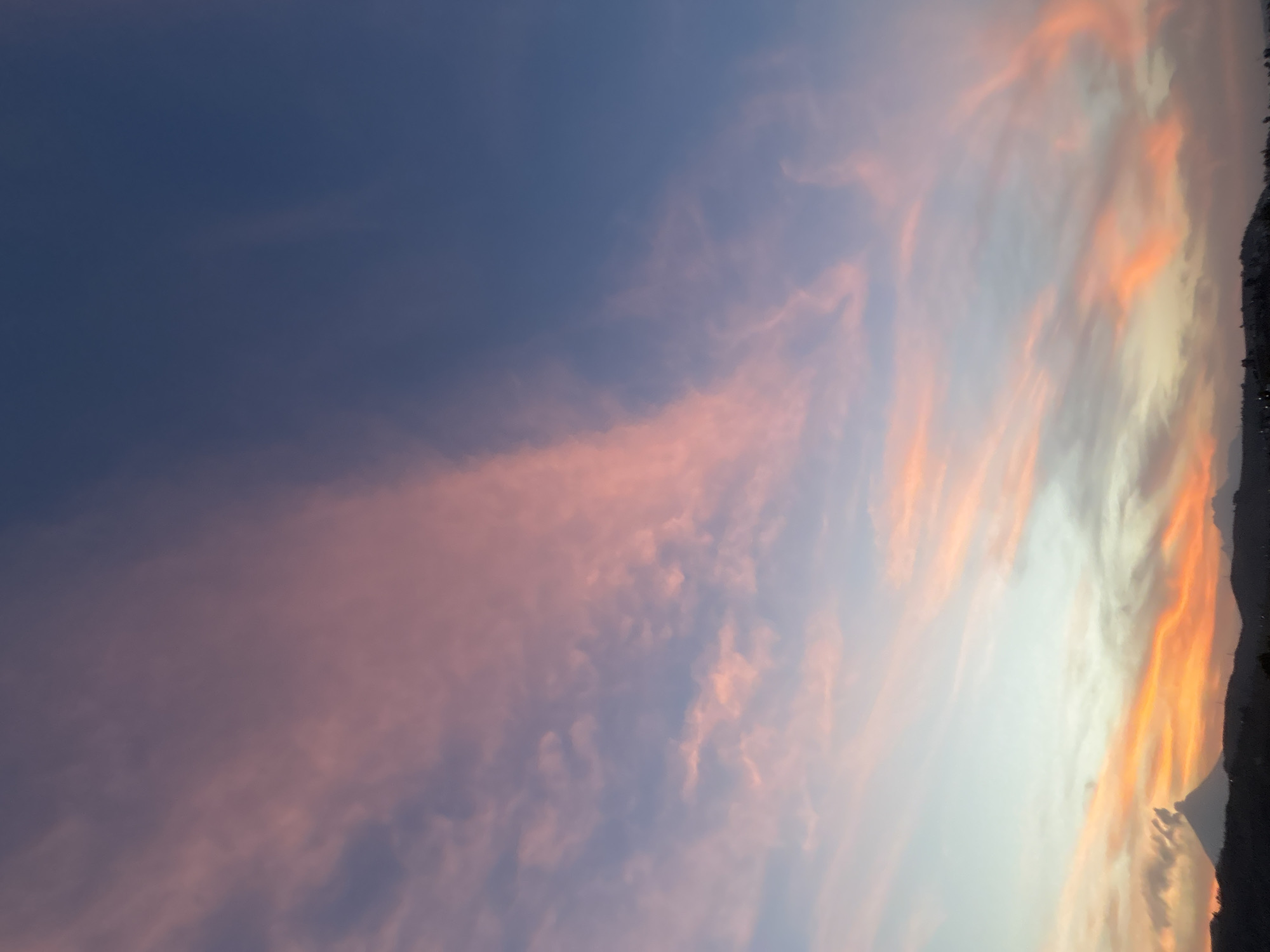 This media file captures a stunning sunset or sunrise sky over Santa Clara Ocoyucan, Mexico, presenting a vivid spectrum of colors. The sky transitions from a deep, muted blue and purplish hue on the left to brilliant shades of pink, orange, and yellow on the right, indicating the proximity of the sun to the horizon.
Wispy, scattered clouds are illuminated by the natural light, appearing in various tones of soft pinks, warm oranges, and hints of yellow, particularly concentrated on the right side of the frame. The clouds create dynamic patterns against the lighter parts of the sky.
Along the far right edge of the image, a dark, jagged silhouette of what appears to be mountains or hills is visible, grounding the expansive sky view. The atmosphere suggests a clear day with scattered clouds, ideal for a dramatic display of natural light. There are no visible people, objects, or identifiable activities taking place, and no discernible text. The overall scene evokes a sense of natural beauty and tranquility at either dawn or dusk.