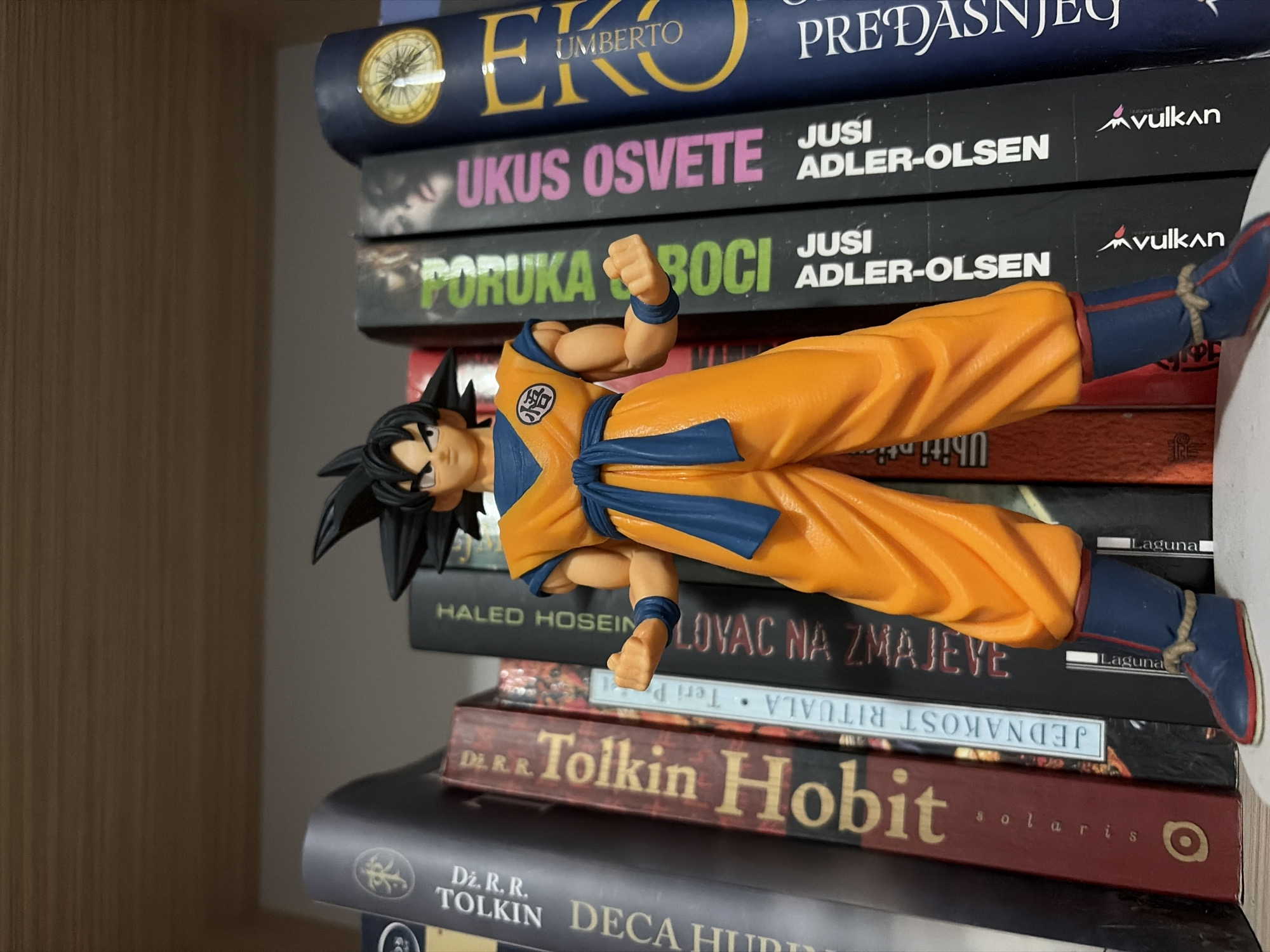 A Goku action figure from Dragon Ball is playfully displayed horizontally against a stack of books, creating the illusion of flight or levitation. The figure, dressed in its iconic orange gi with blue details, has one foot lightly resting on the book stack while the other is extended. Its right arm is bent with a clenched fist, and the left arm is extended forward. The scene is set indoors, likely on a wooden bookshelf, visible on the left side of the frame.
The books, mostly translated into Serbian, include notable titles and authors:
- **Umberto Eco** with "PREDASNJEG" (suggesting a historical or older work).
- Two thrillers by **Jussi Adler-Olsen**: "UKUS OSVETE" (Taste of Revenge) and "PORUKA" (likely part of "Poruka u boci" - Message in a Bottle), both published by Vulkan.
- **Khaled Hosseini's** "LOVAC NA ZMAJEVE" (The Kite Runner), published by Laguna.
- **J. R. R. Tolkien's** "Hobit" (The Hobbit), published by Solaris.
- Another Tolkien work, "DECA HURIN" (The Children of Húrin).
The arrangement suggests a personal collection, creatively merging pop culture with literature. The image is well-lit, indicative of an indoor daytime setting.